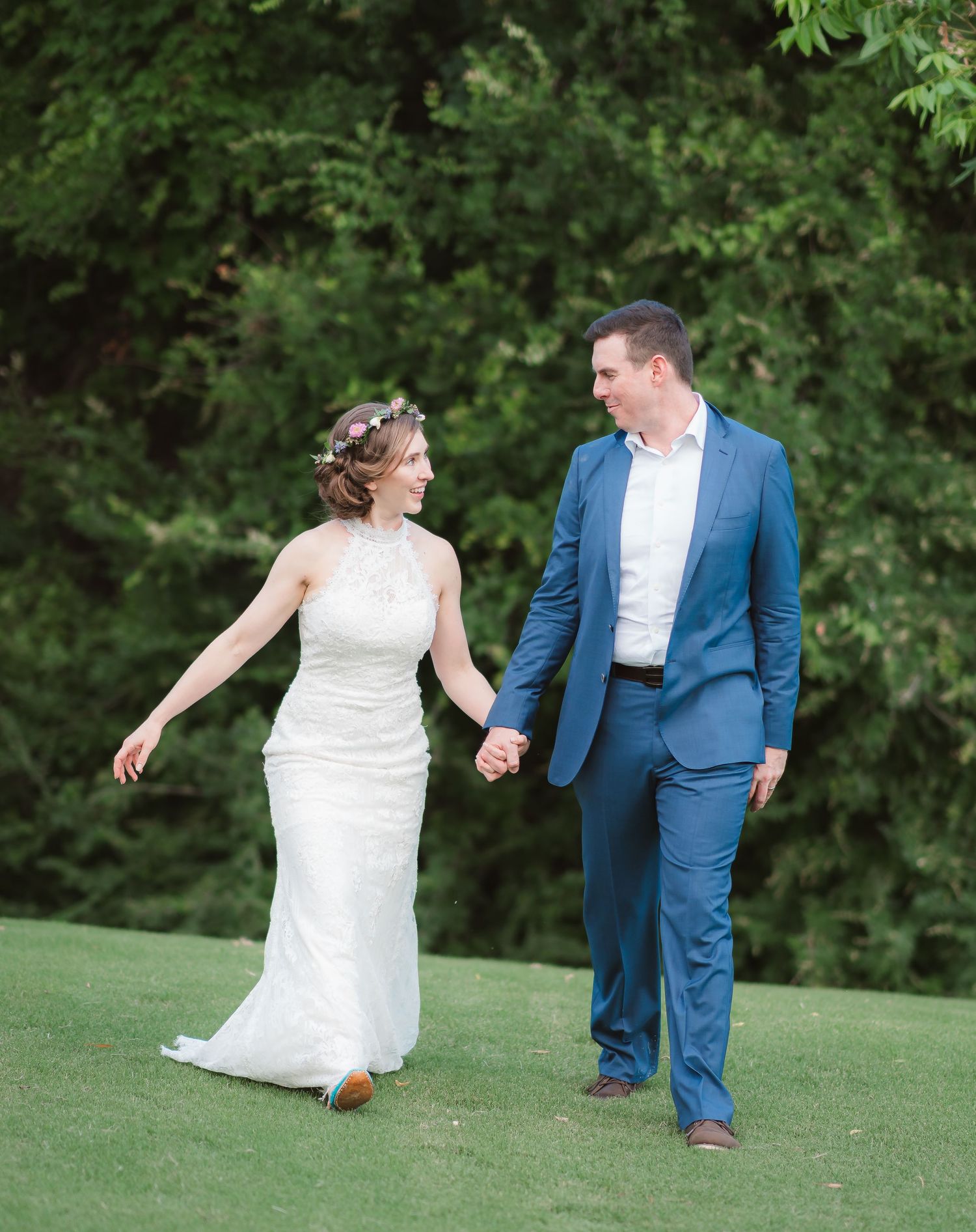 A couple in wedding attire playfully runs and dances together on a lush green lawn surrounded by trees.