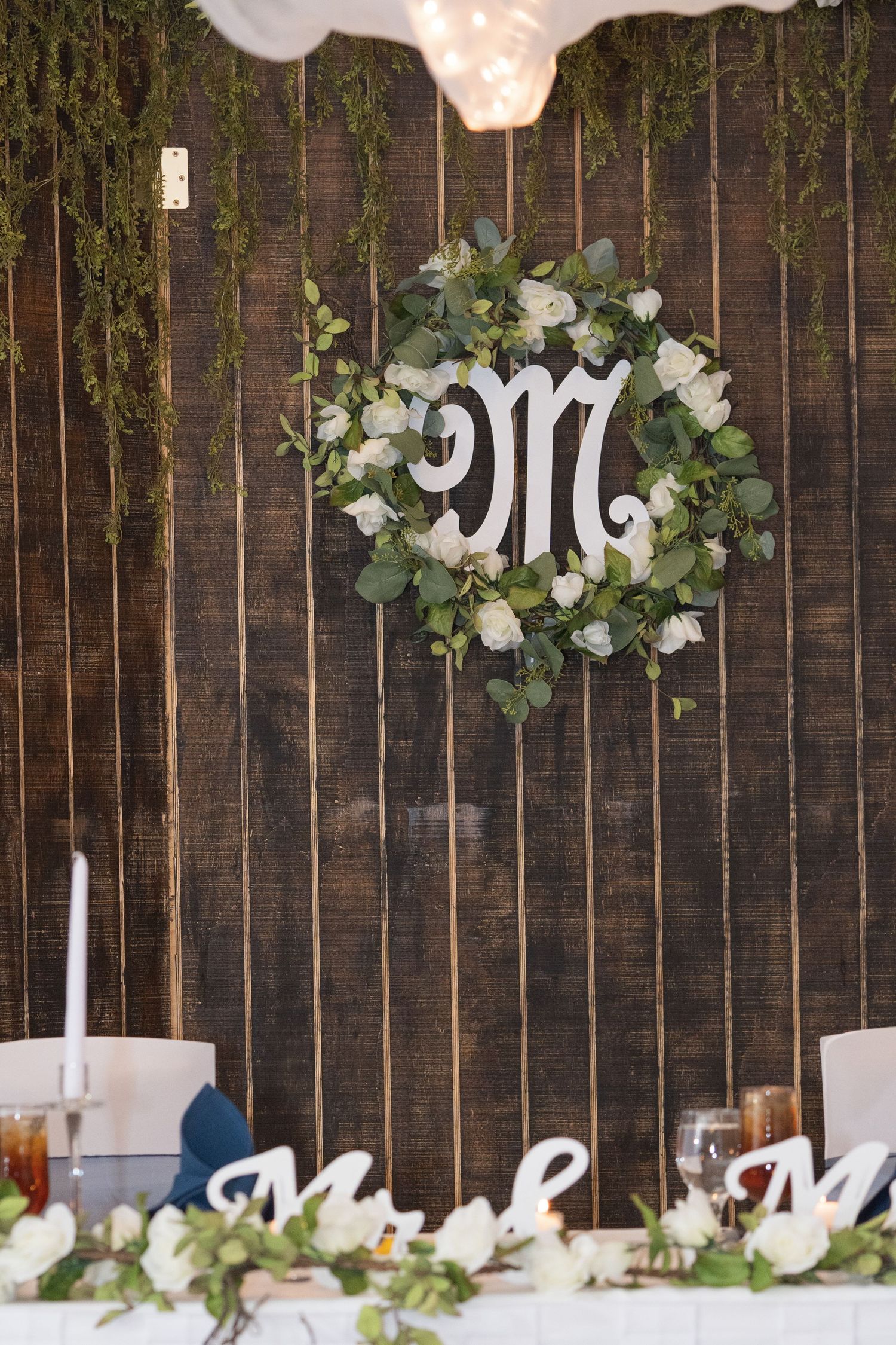 Wedding reception decor featuring white floral arrangements and greenery against a rustic wooden wall backdrop.