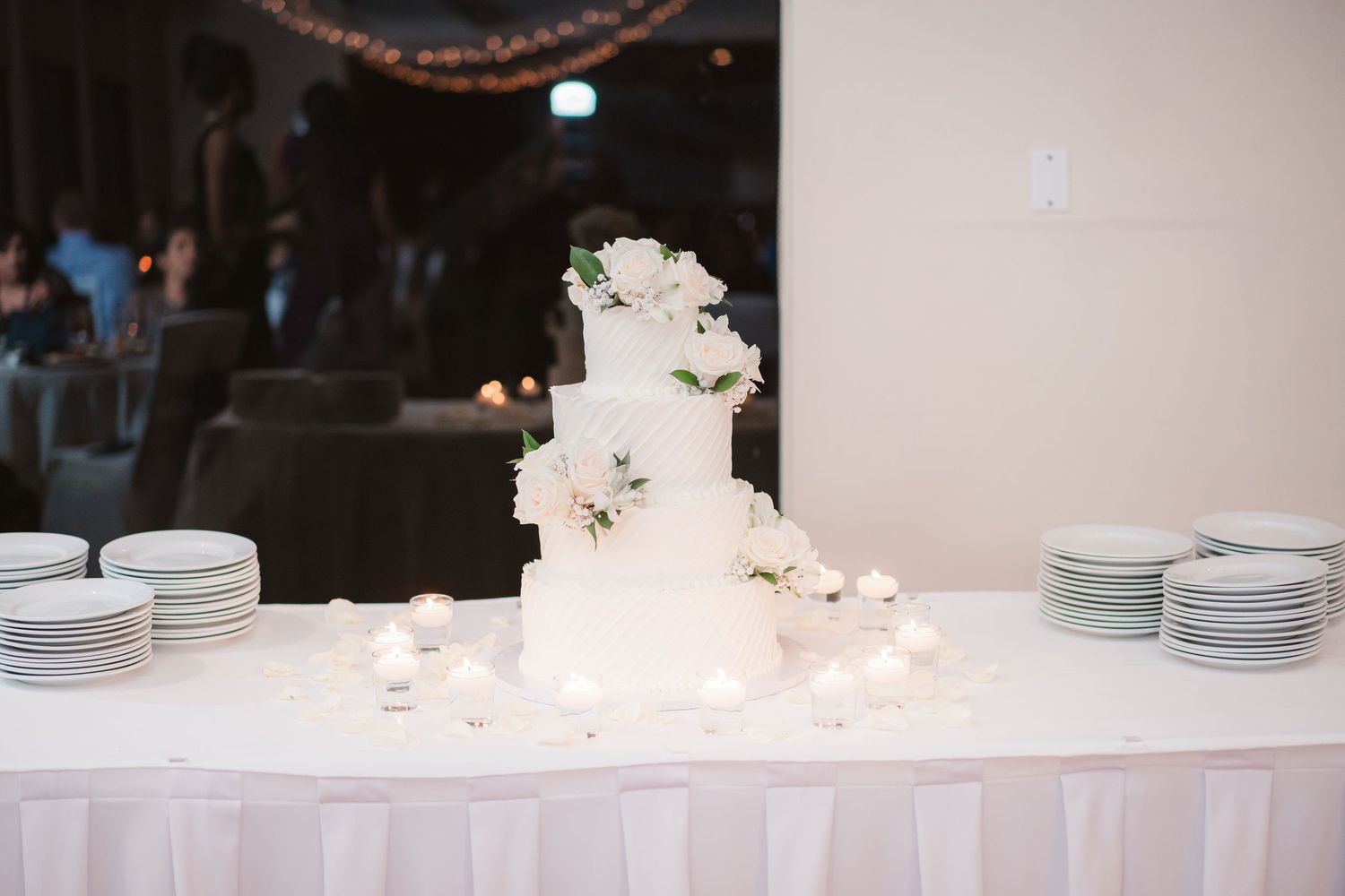 A white wedding cake display table with elegant place settings and soft glowing candle lights along the edge.