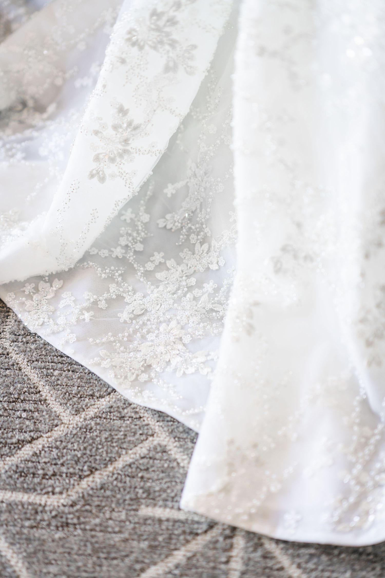 Close-up detail of delicate white lace embroidery on wedding dress fabric against gray carpet.
