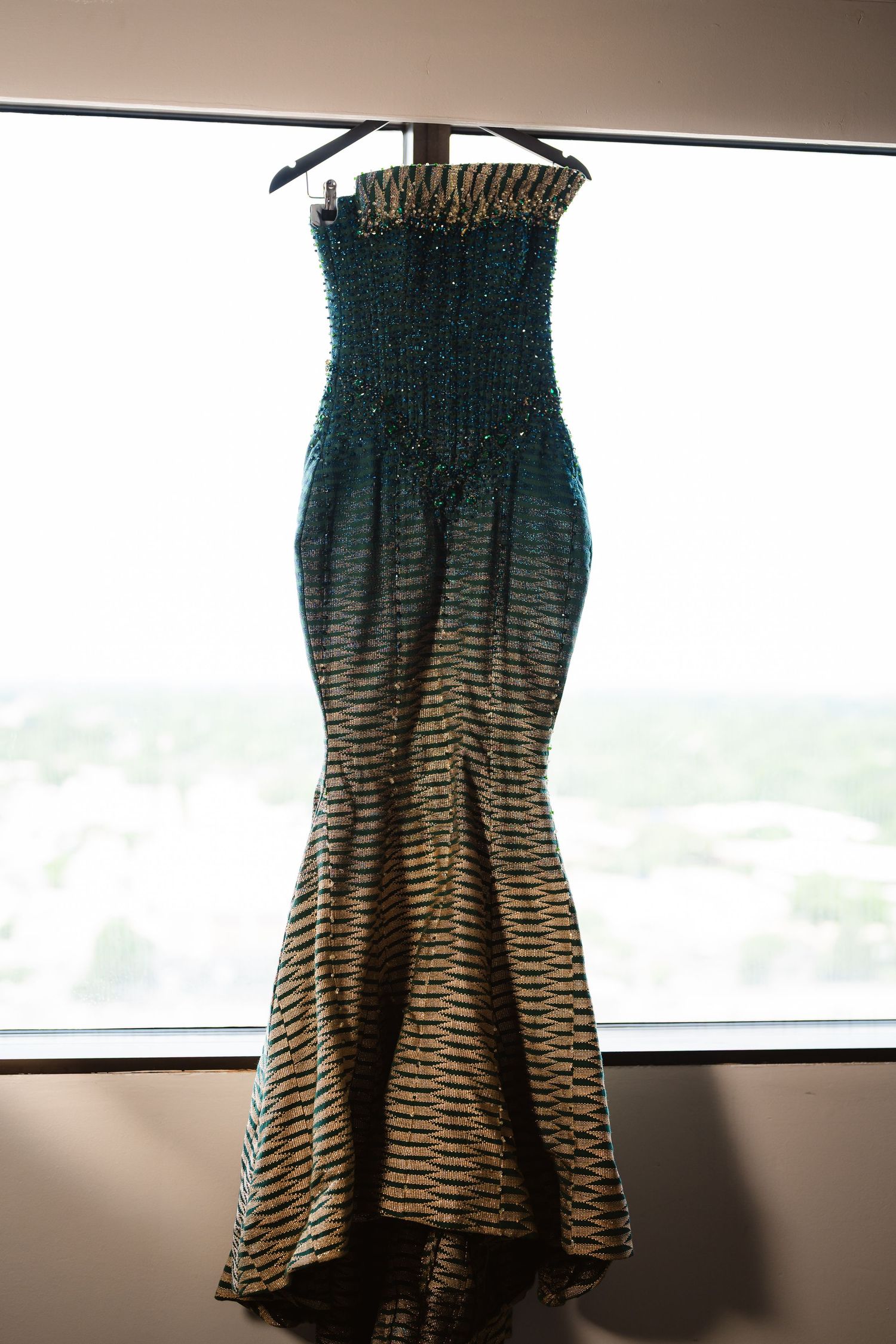 A teal and brown ombre dress with smocked bodice hangs against a window with natural lighting.