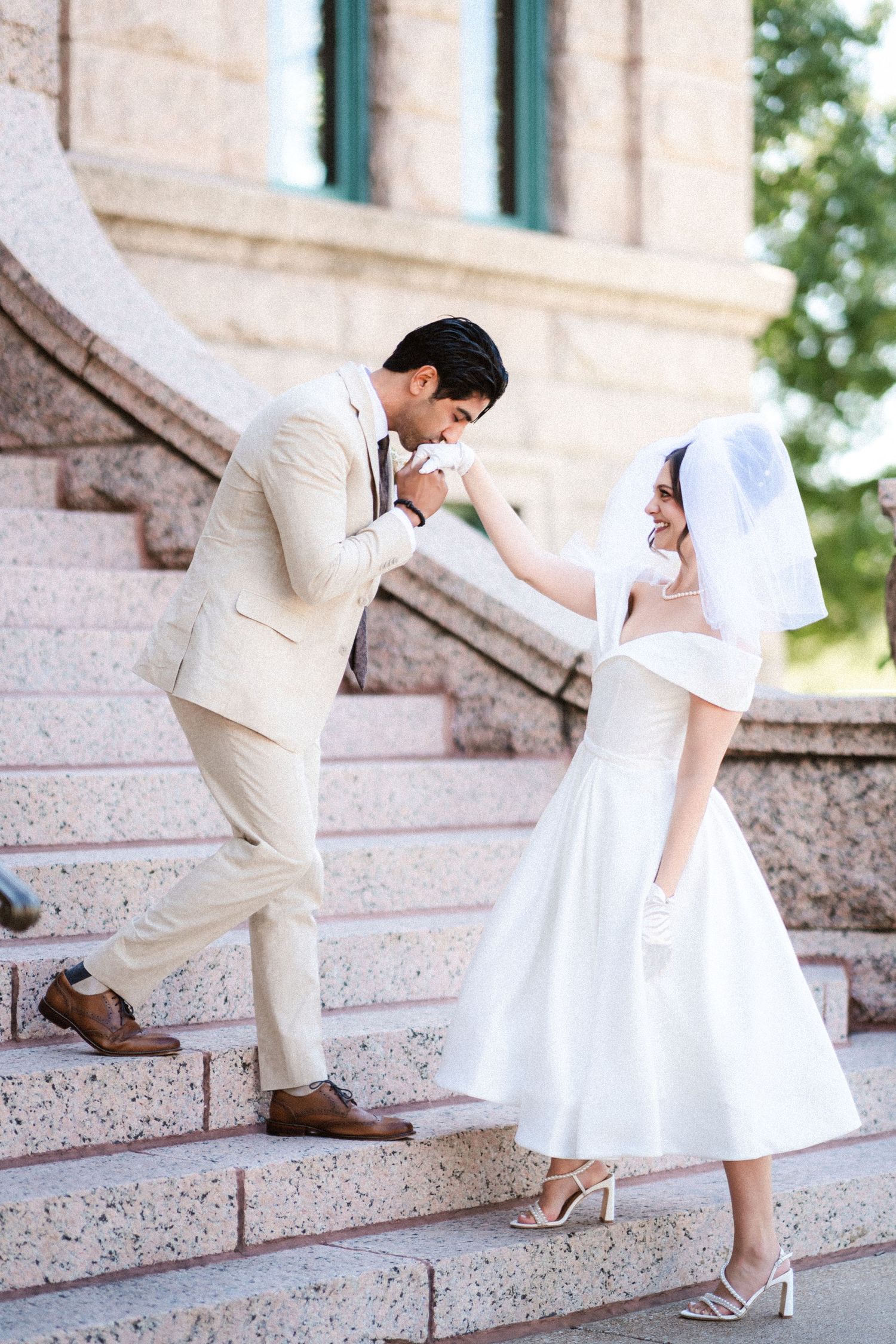 A couple in coordinating white and beige outfits share a tender moment on outdoor stone steps.