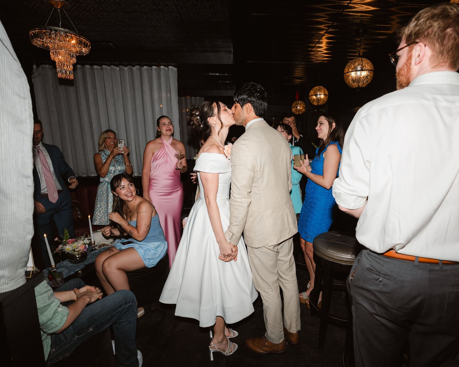 Wedding reception party scene with guests dancing and celebrating in a dimly lit venue.
