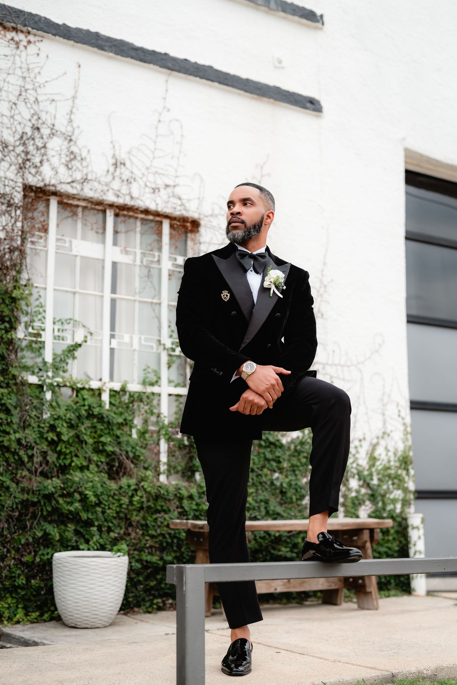 Groom in formal attire sits on wooden bench against white building with potted plants and windows.
