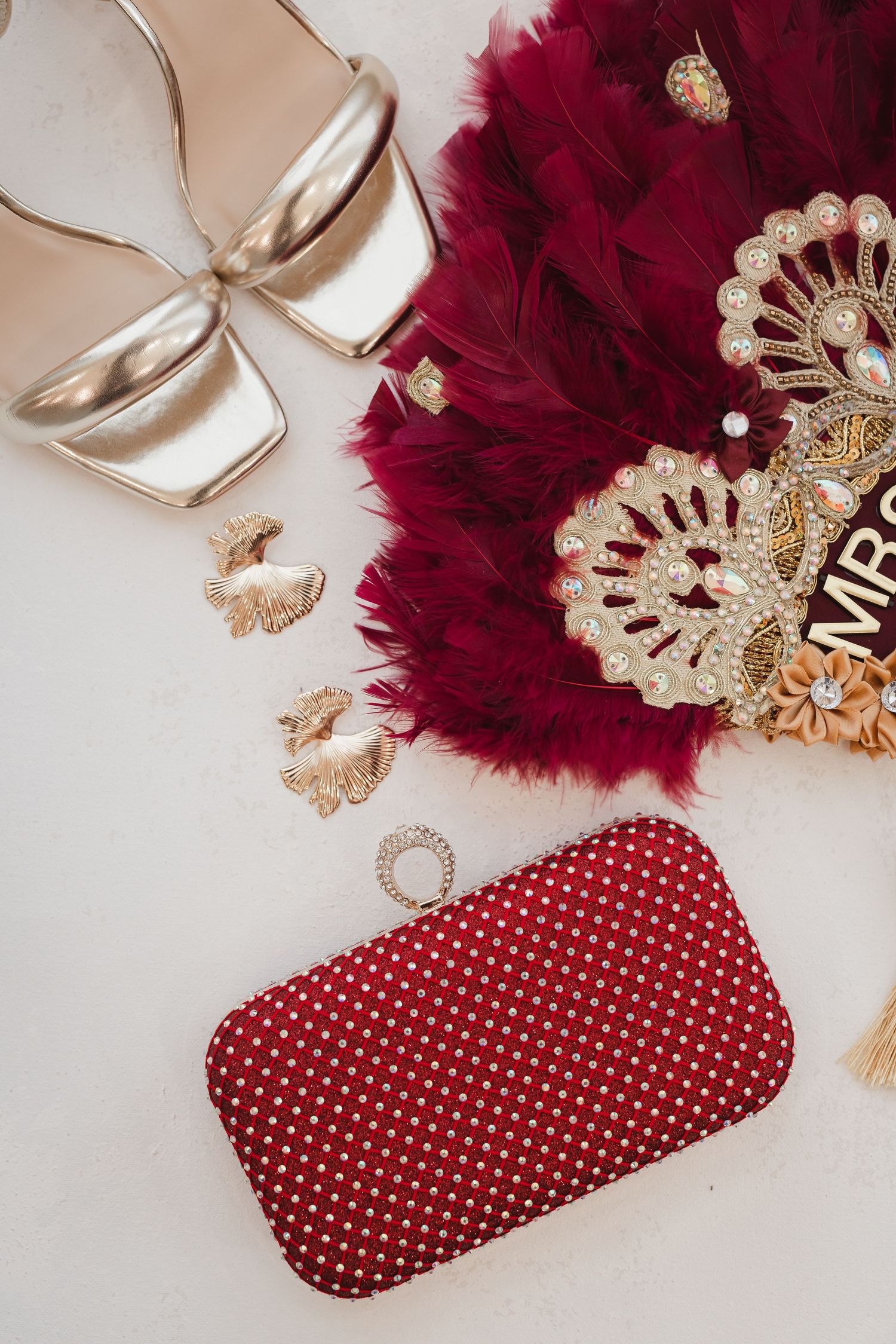 Burgundy feather fan with gold lace trim and polka dot clutch arranged on white surface with decorative elements.