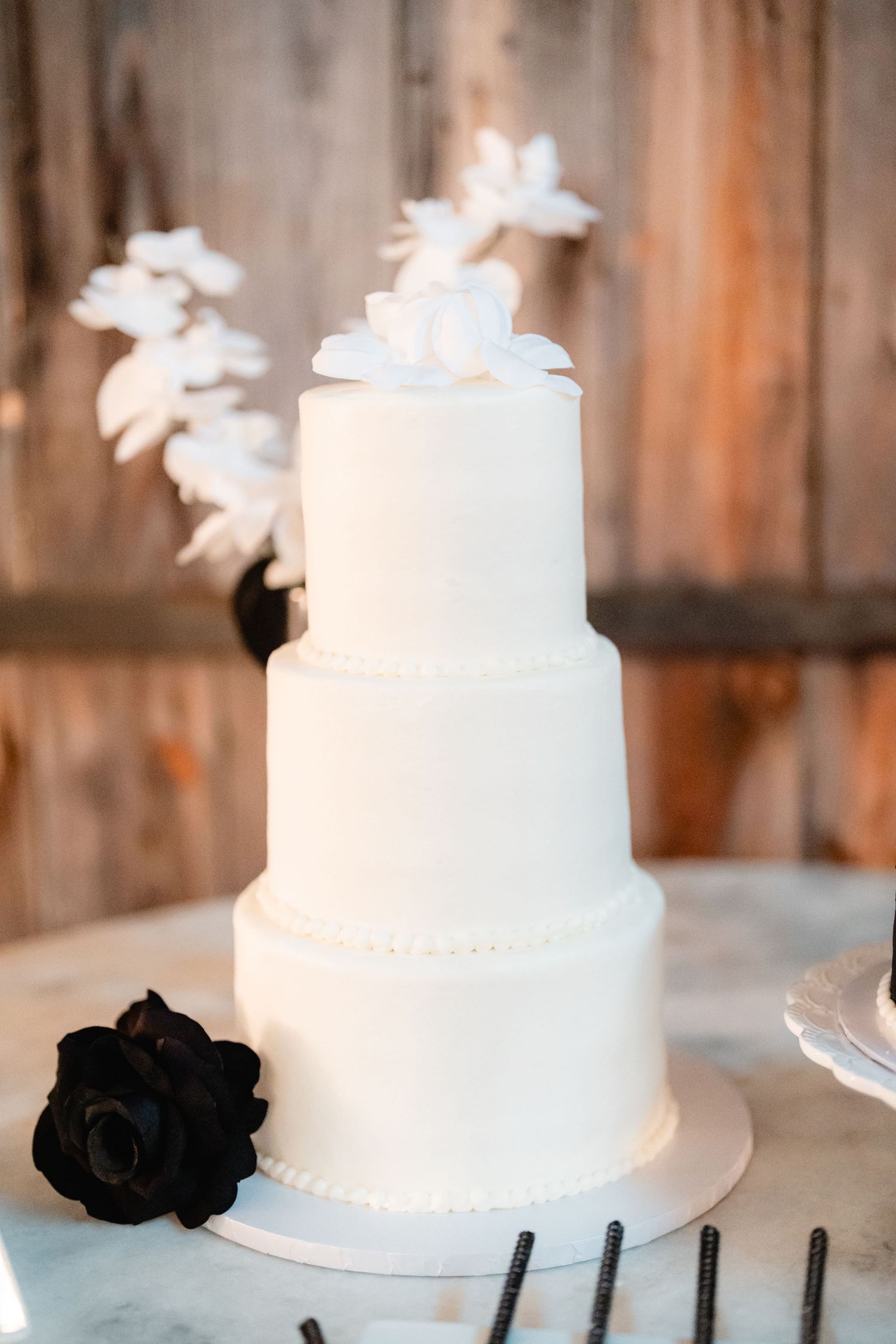 White tiered wedding cake with black rose accent and floral topper on rustic wooden background.