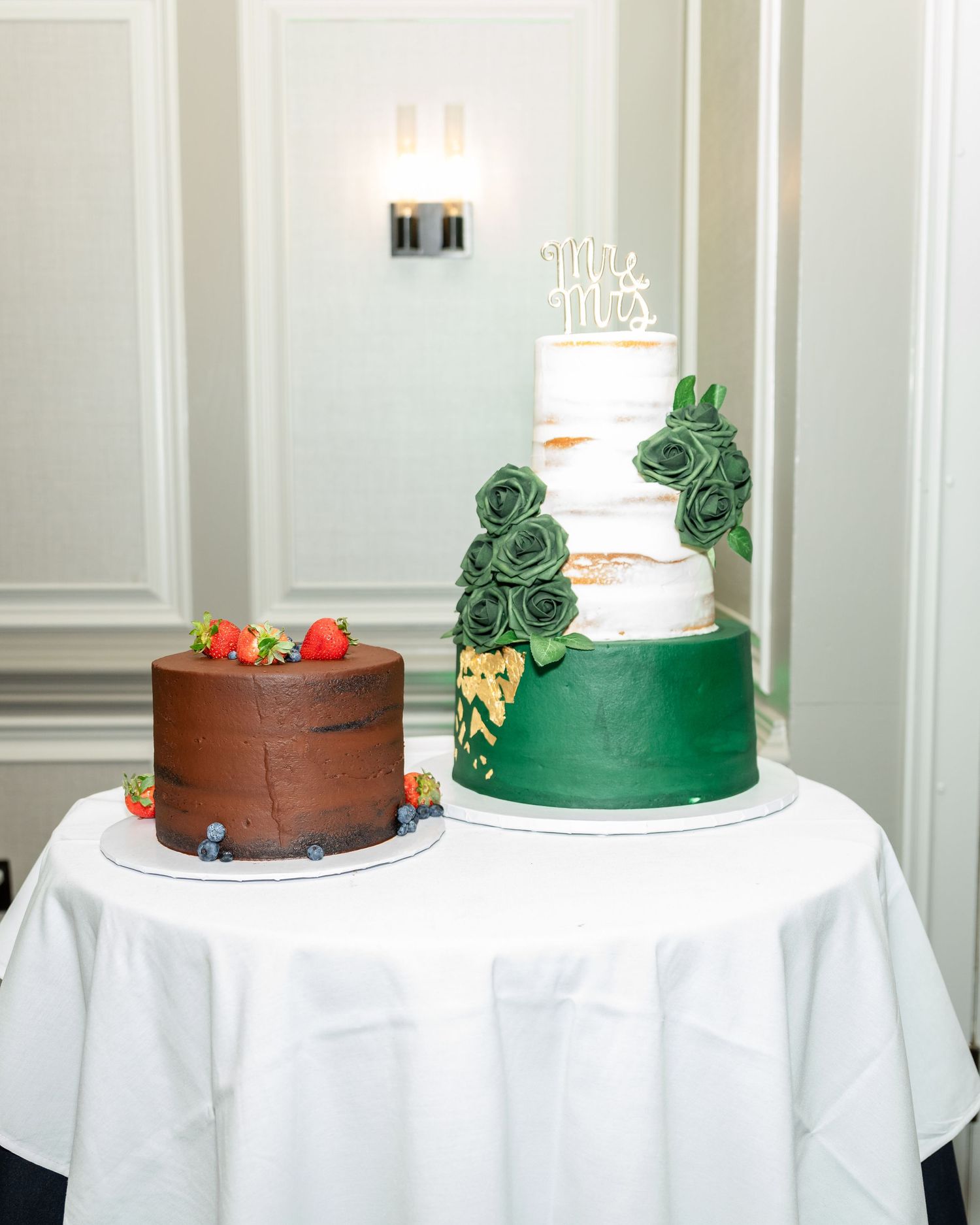 Two wedding cakes on white tablecloth, one brown and one white with green floral decorations.