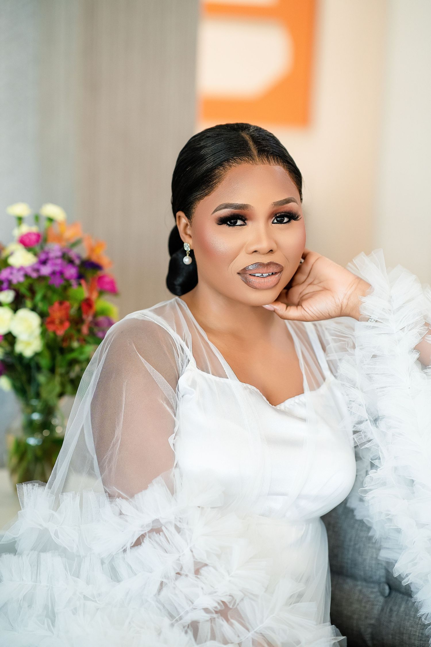 A bride in a romantic white tulle gown with sheer sleeves poses elegantly next to colorful floral arrangements.