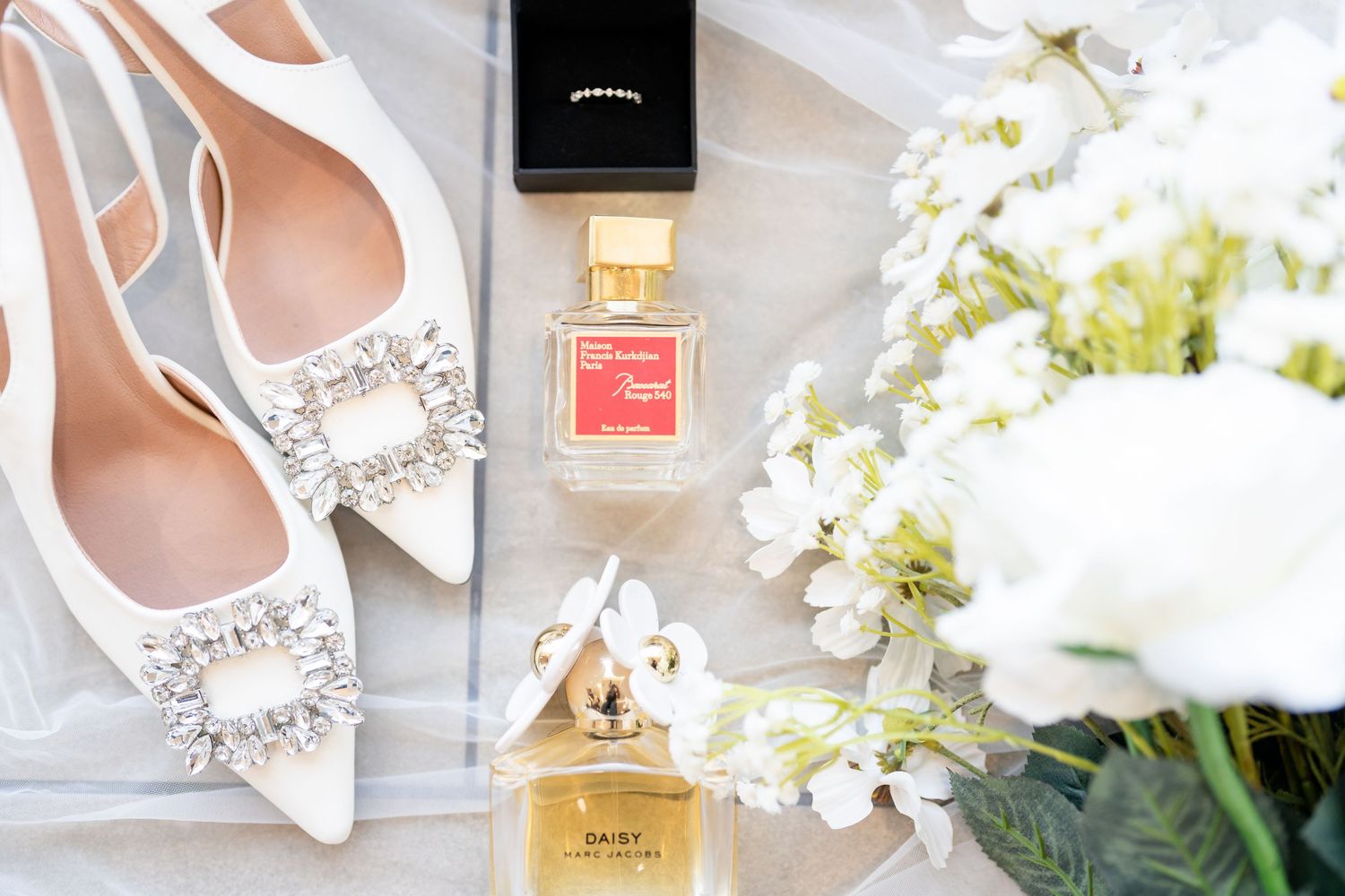 Elegant bridal details including white shoes, perfume bottle, jewelry and delicate white flowers arranged on light surface.