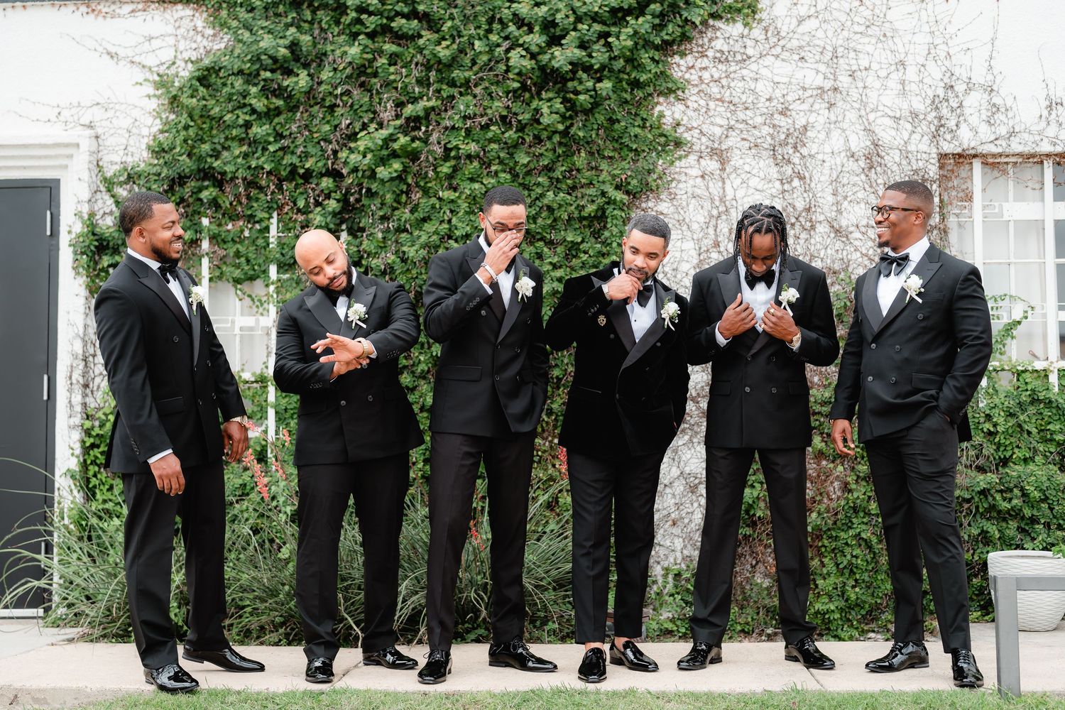 Groomsmen in black tuxedos sharing a candid moment while standing against an ivy-covered wall.