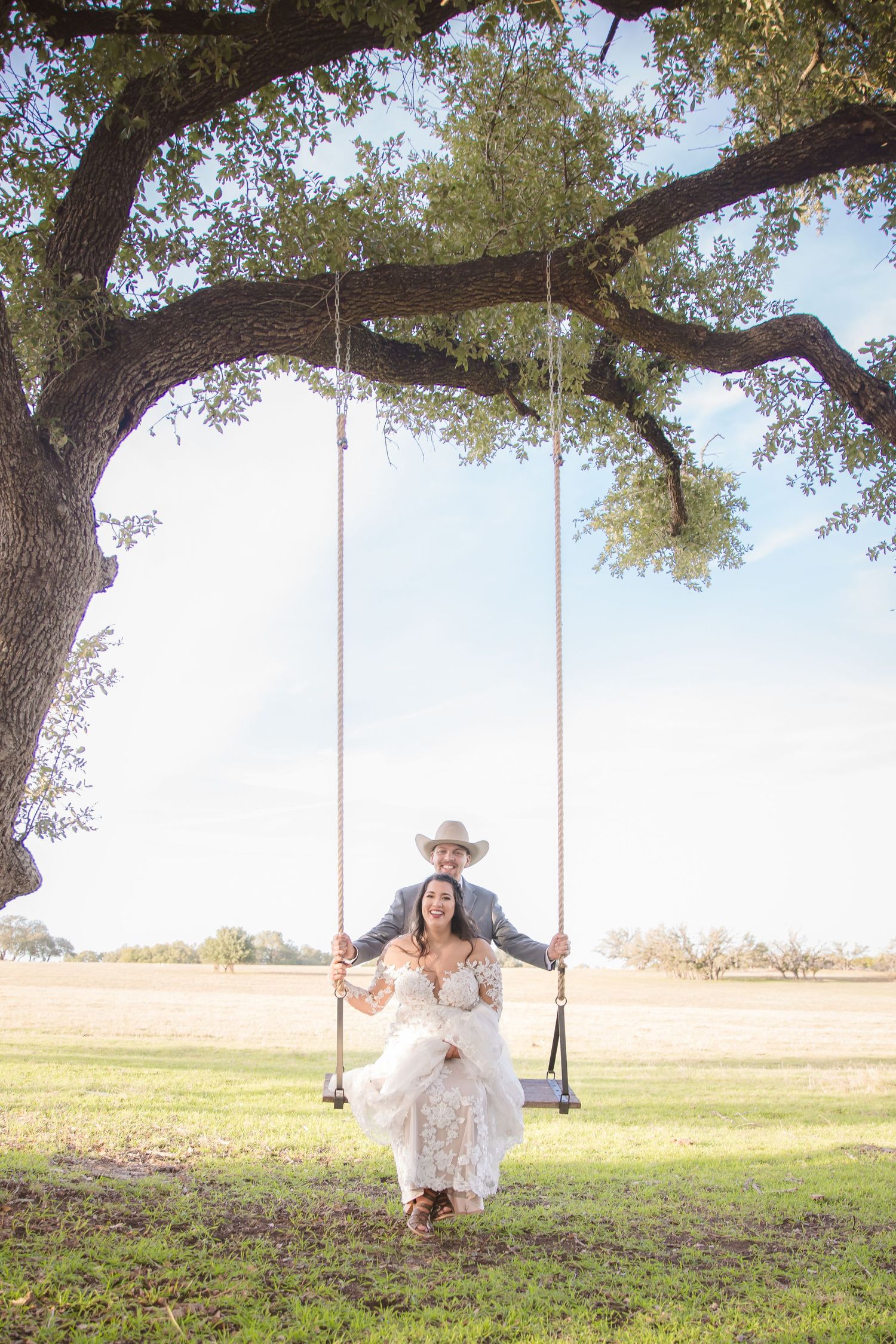 A couple in wedding attire enjoys swinging together under a large oak tree in a pastoral setting.