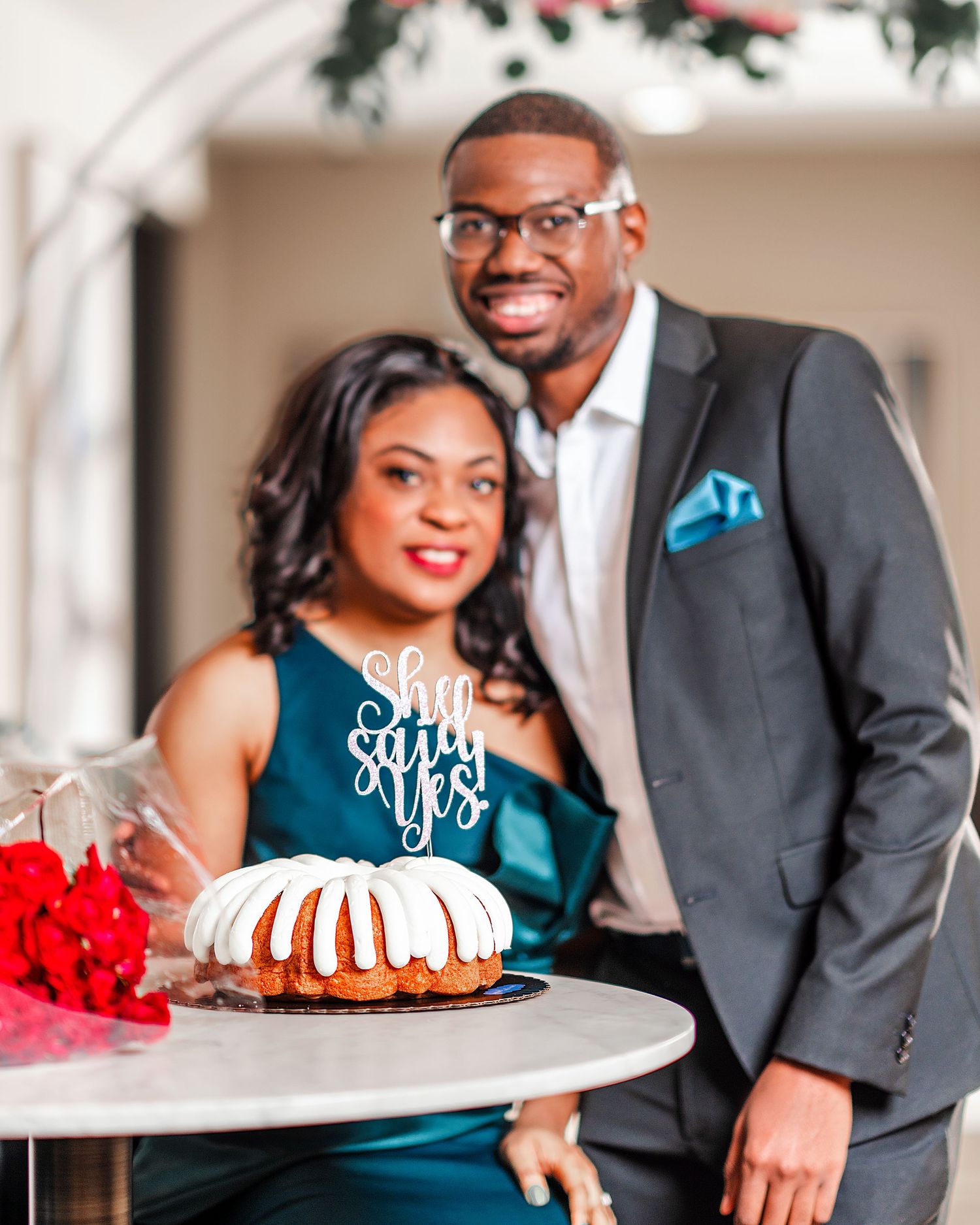 A romantic couple poses with a decorated cake during what appears to be an engagement or celebration photoshoot.