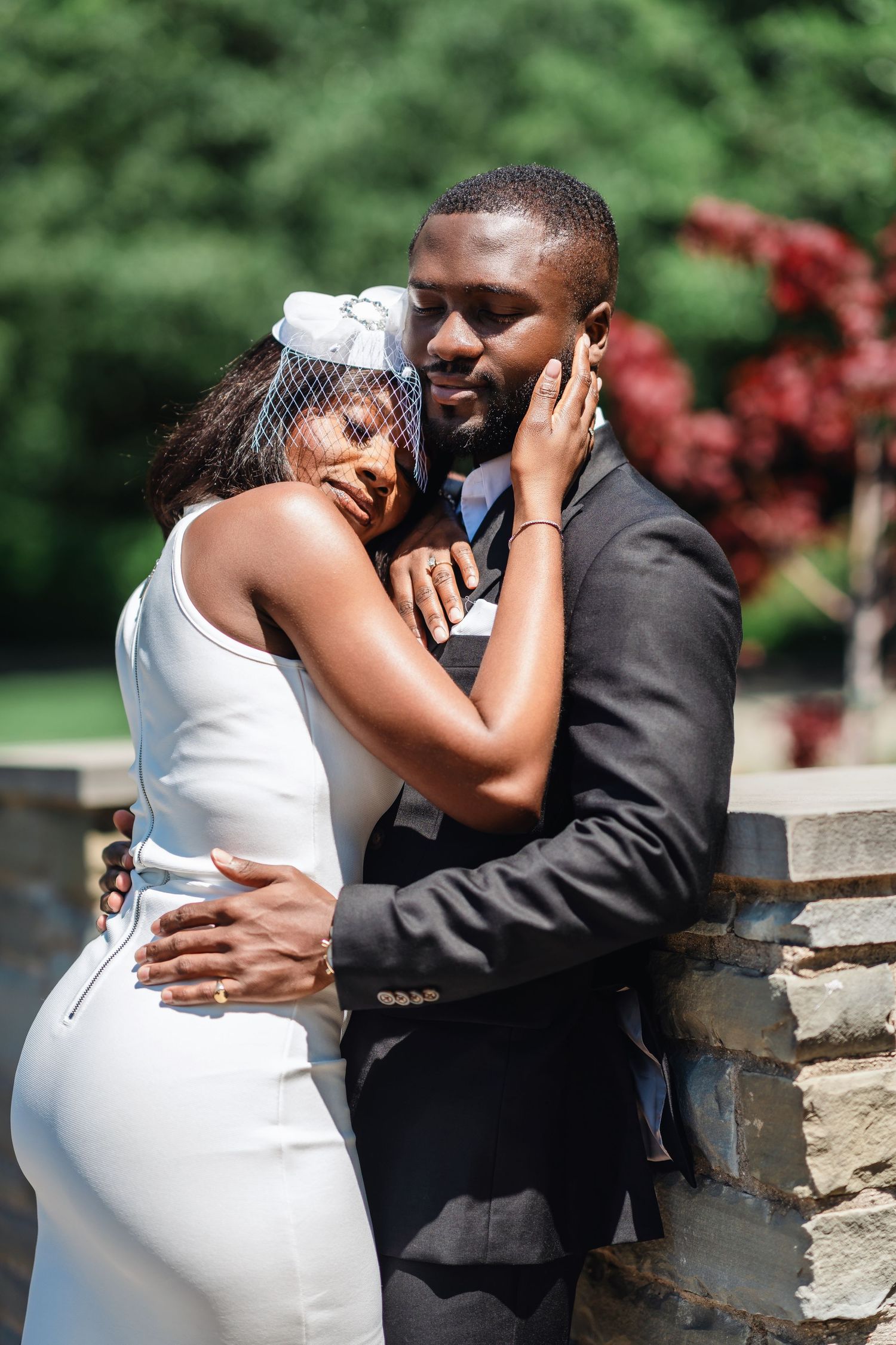 A romantic wedding couple embraces against a lush green backdrop, the bride in white and groom in black suit.