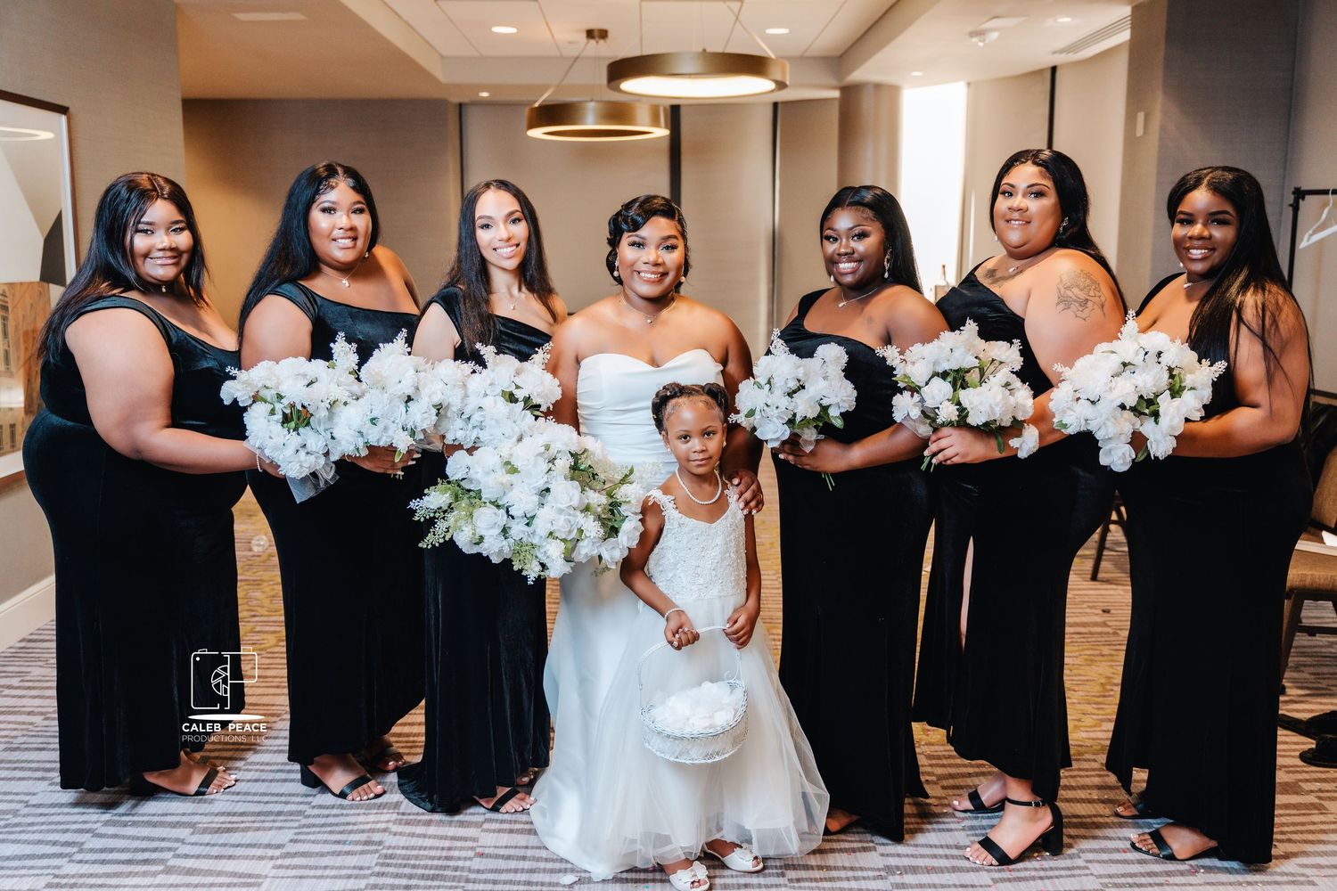 Wedding party lined up wearing black dresses and holding white floral bouquets in an elegant venue.
