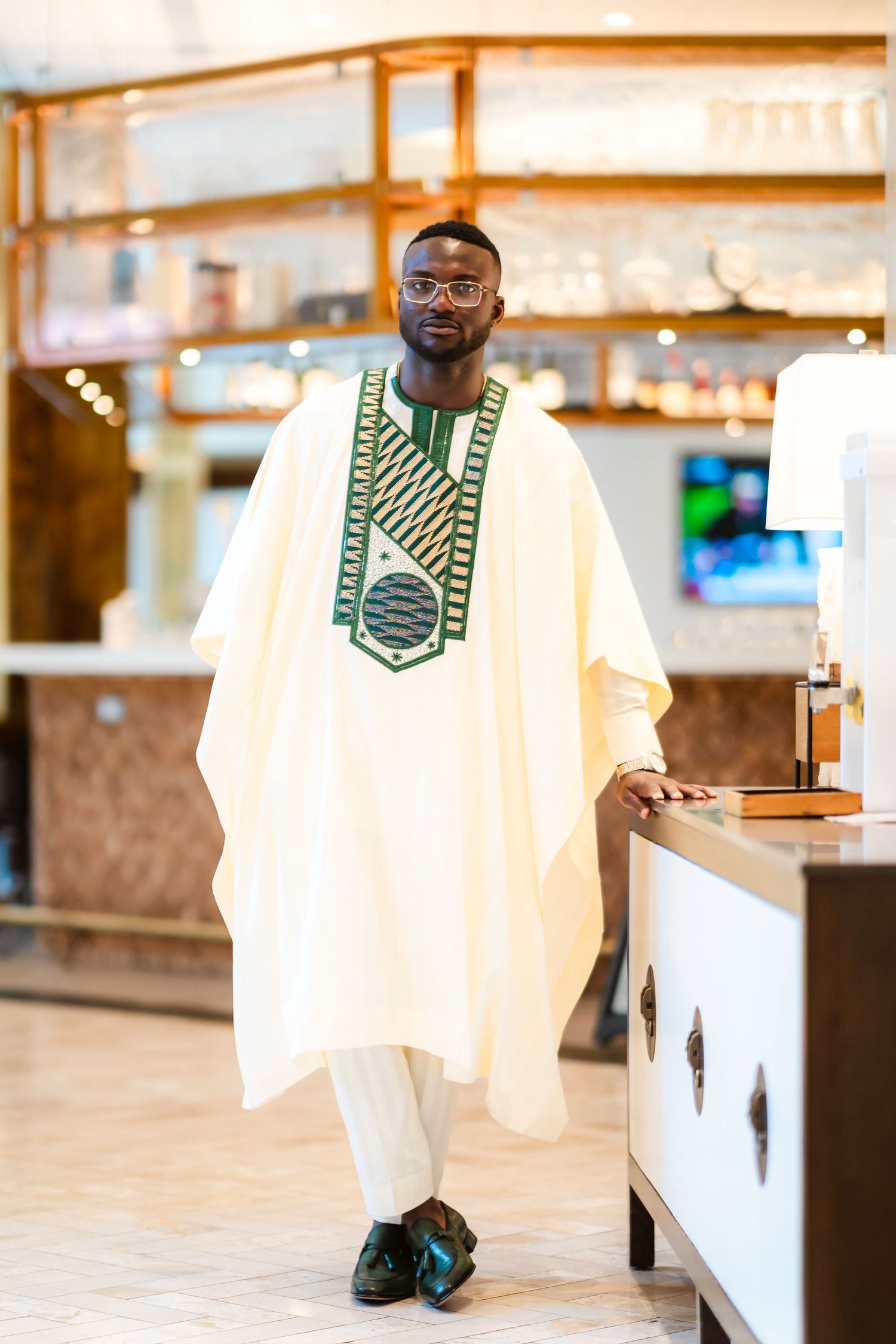 A person wearing a white traditional agbada with green embroidery stands in an elegant indoor setting.