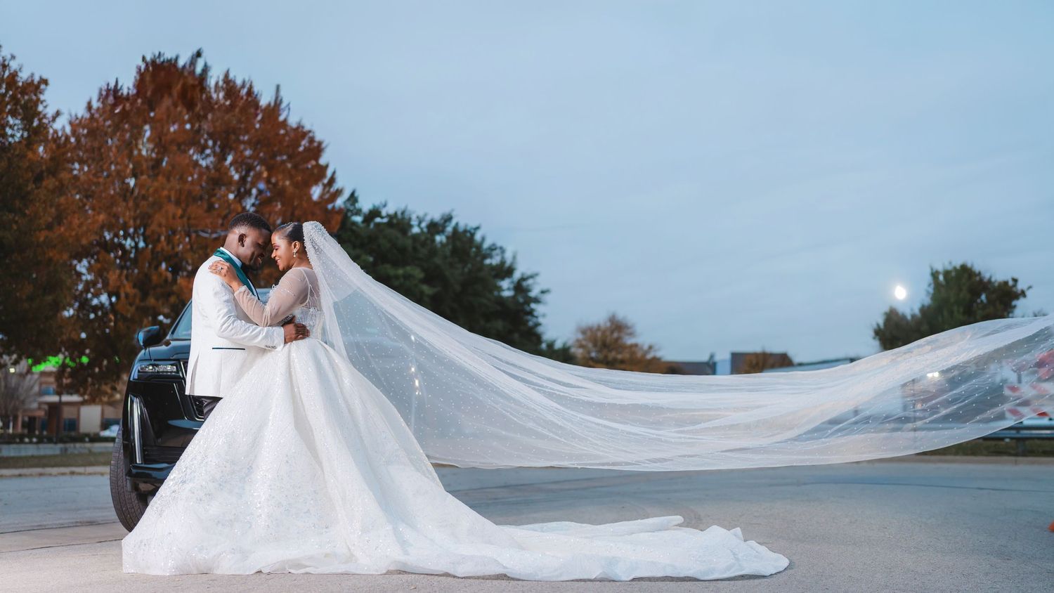 A long white wedding veil flows in the wind against a blue sky with autumn trees.