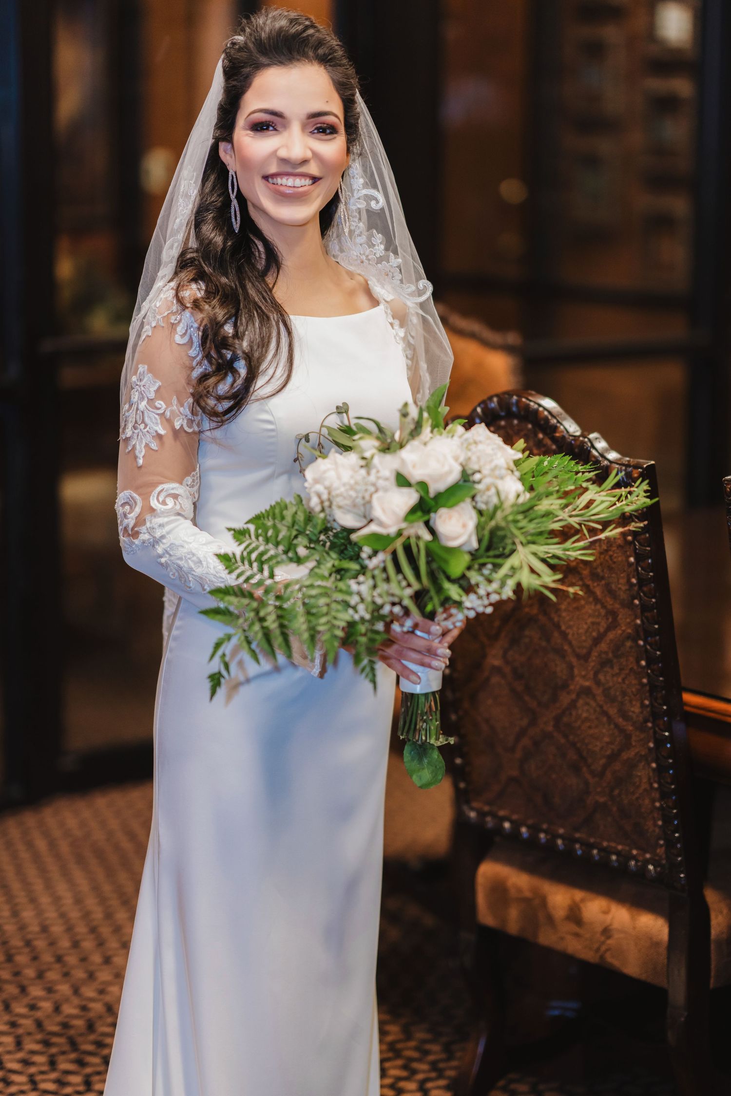 Bride in a long-sleeved white wedding dress holds an elegant green and white floral bouquet in an indoor setting.