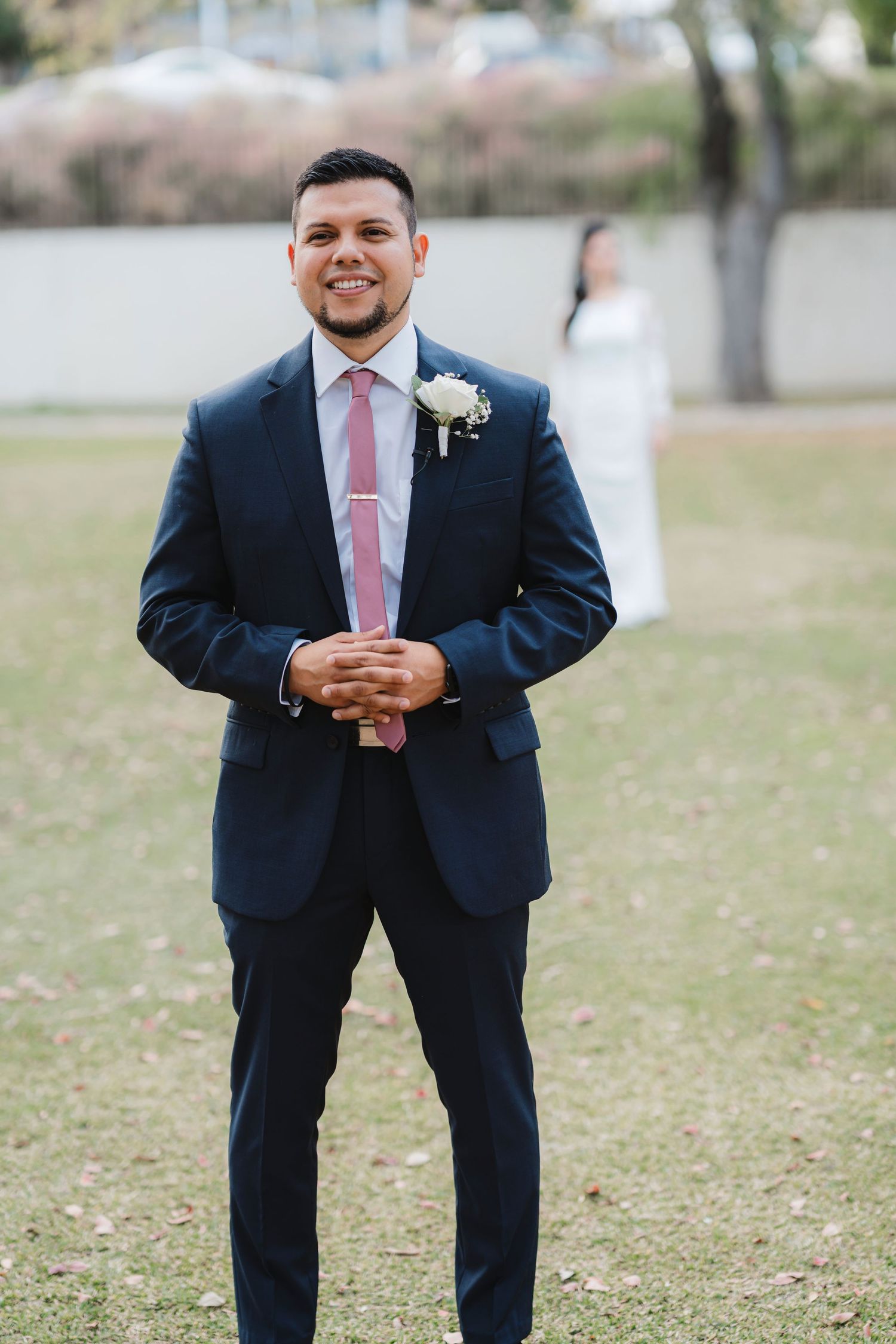 Groom in navy suit with pink tie poses outdoors in a grassy field.