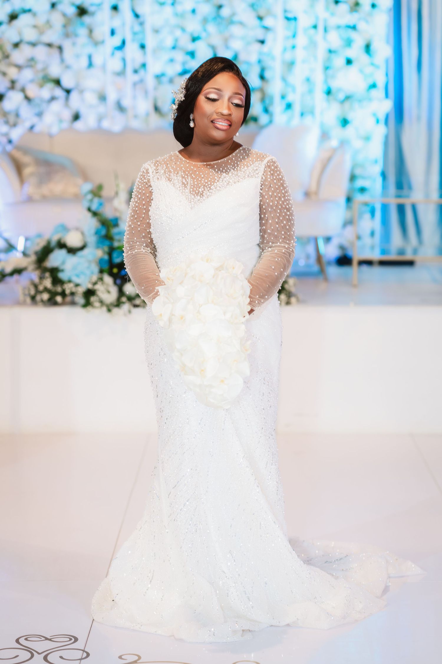 Elegant white wedding dress with sheer polka dot sleeves and floral details against a blue and white backdrop.