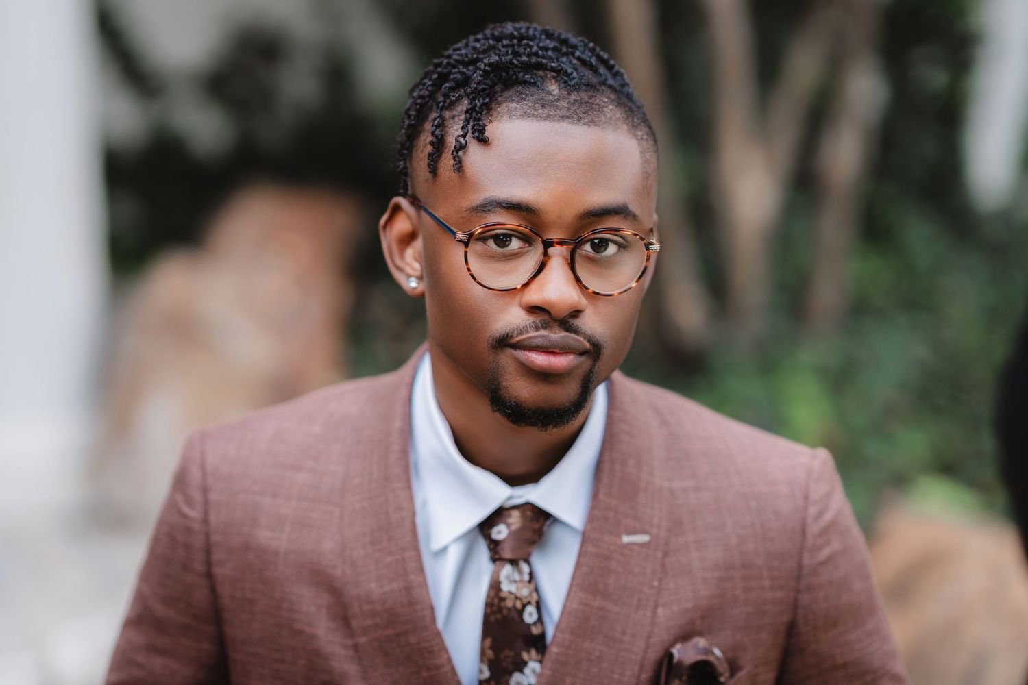 Professional portrait of someone wearing a brown suit, patterned tie, and round glasses in an outdoor setting.