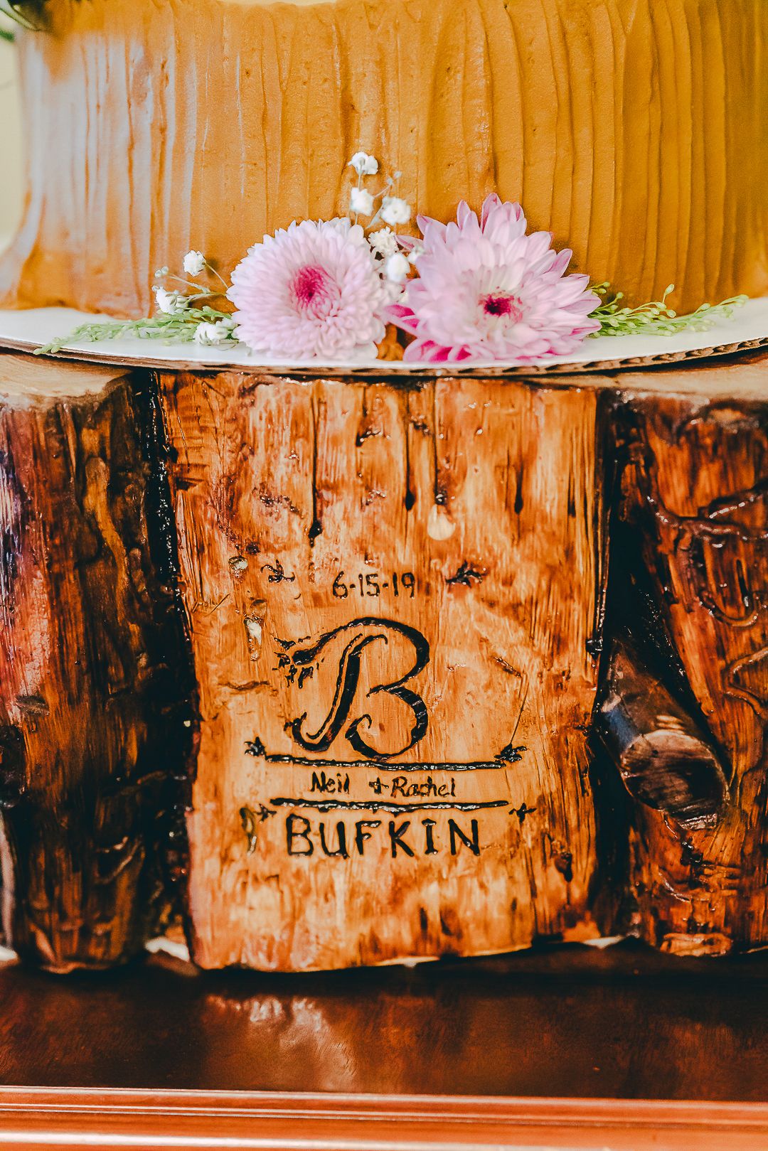 A wooden log decorated with pink gerbera daisies and carved with the name 'Bufkin' serves as a rustic wedding decor piece.