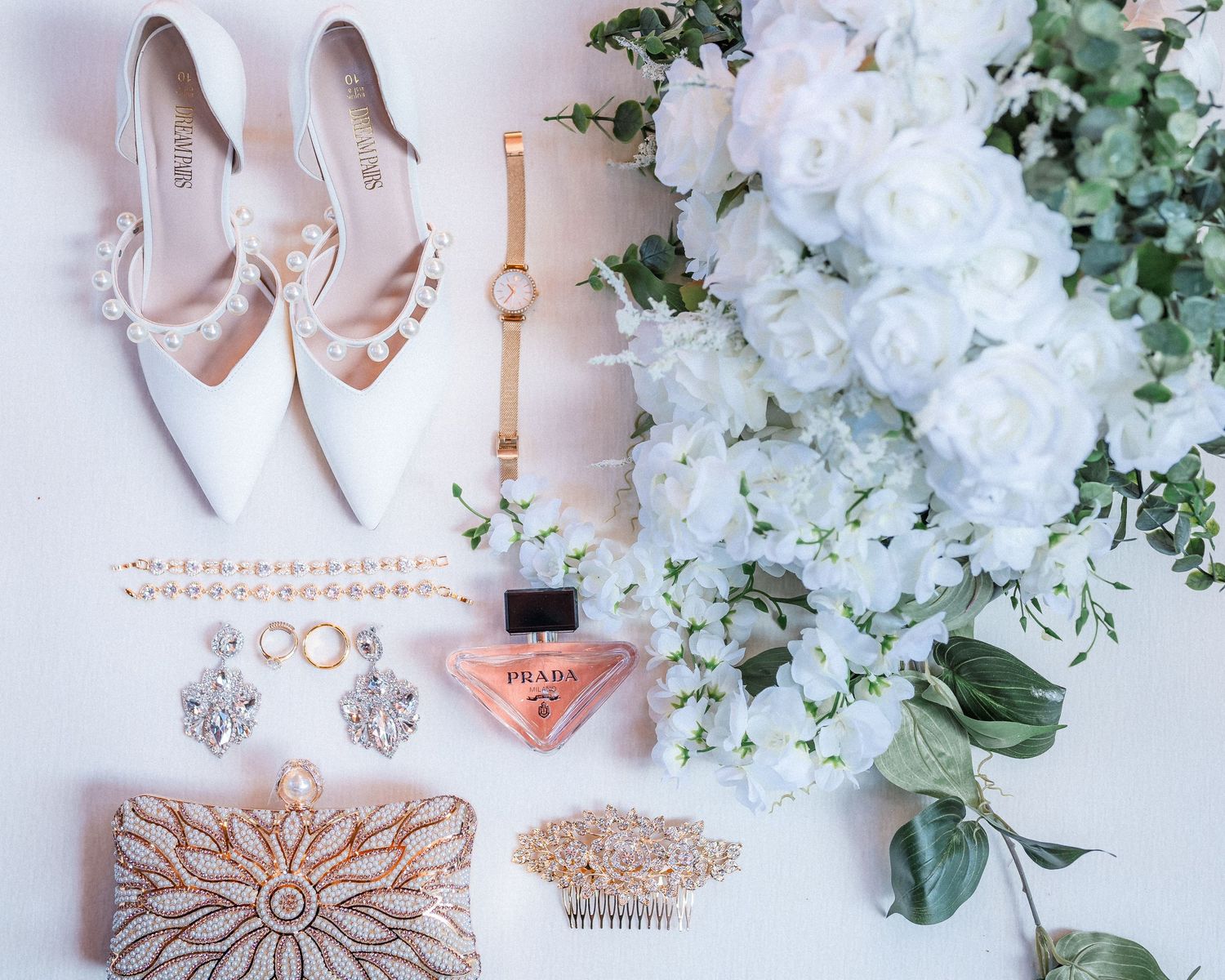Wedding accessories including blush pink heels, jewelry, and white floral arrangements on a light background.