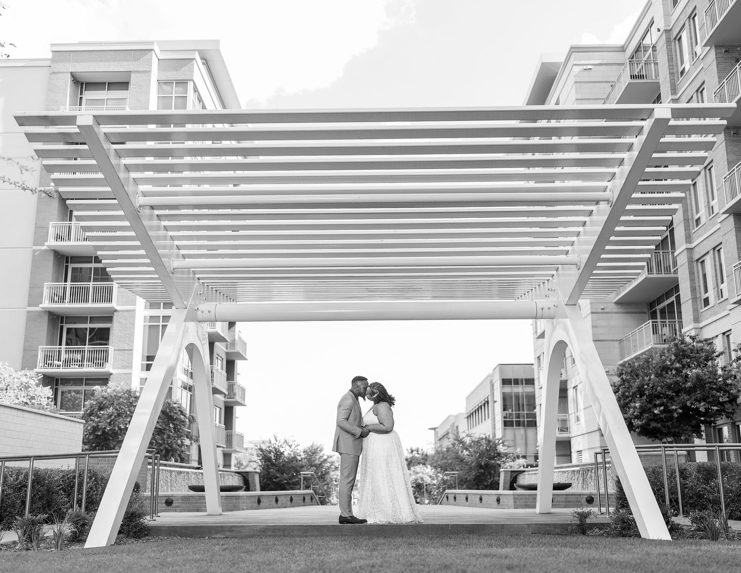 A couple shares a romantic moment under a modern white pergola structure outside an urban building complex.