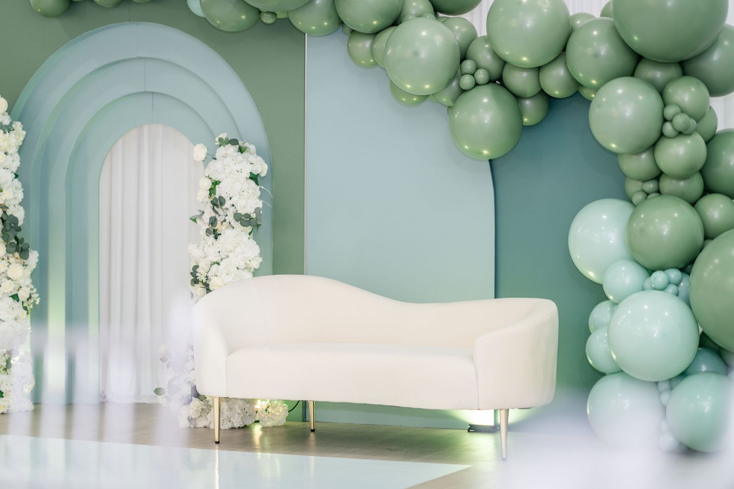 Elegant sage green balloon arch installation with white floral accents decorates an archway entrance.