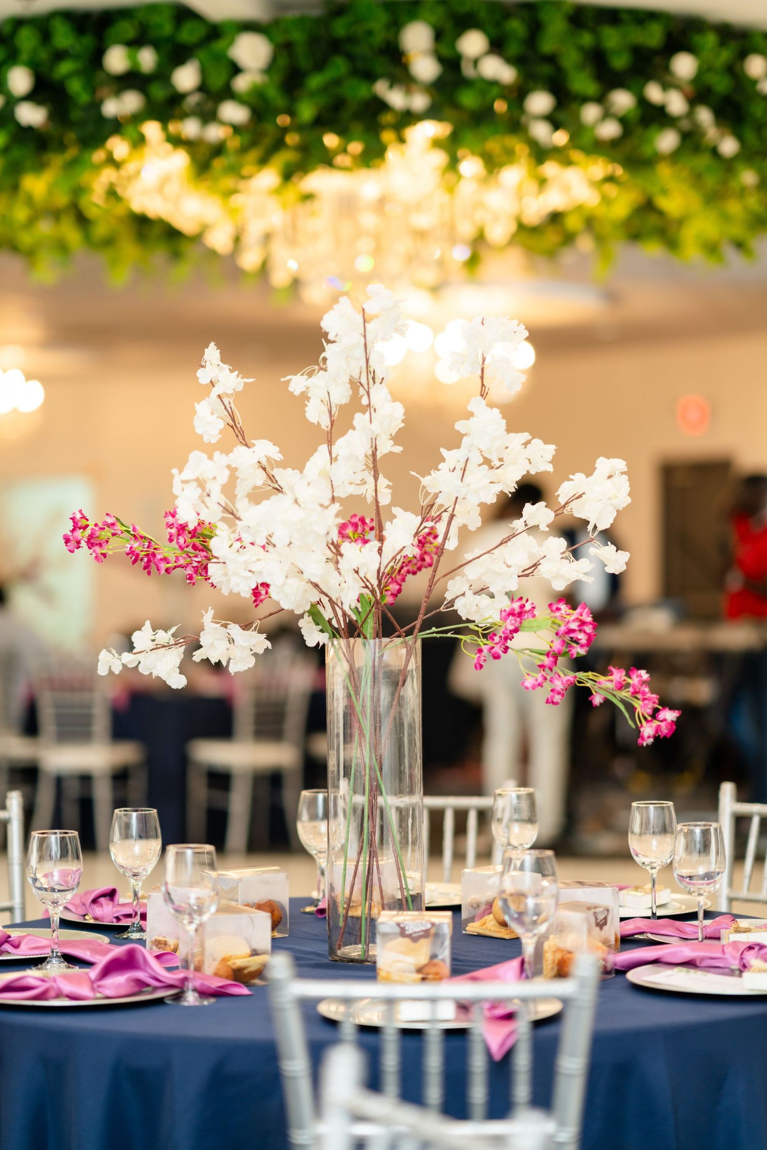Elegant wedding reception table with navy tablecloth and pink orchid centerpiece in tall glass vase under string lights.