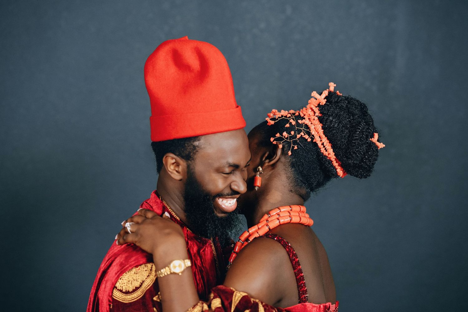 A couple wearing vibrant red and orange traditional African wedding clothing embrace in a studio setting.