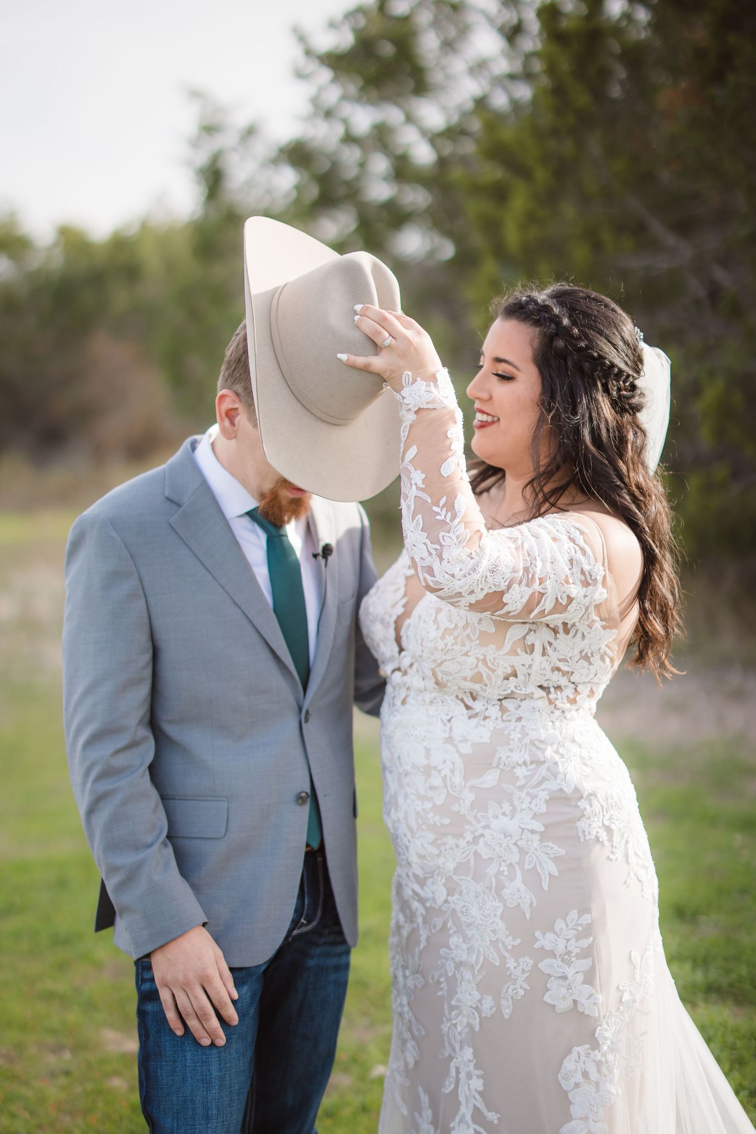 A playful wedding moment captured as the bride lifts the groom's cowboy hat while both laugh in an outdoor setting.