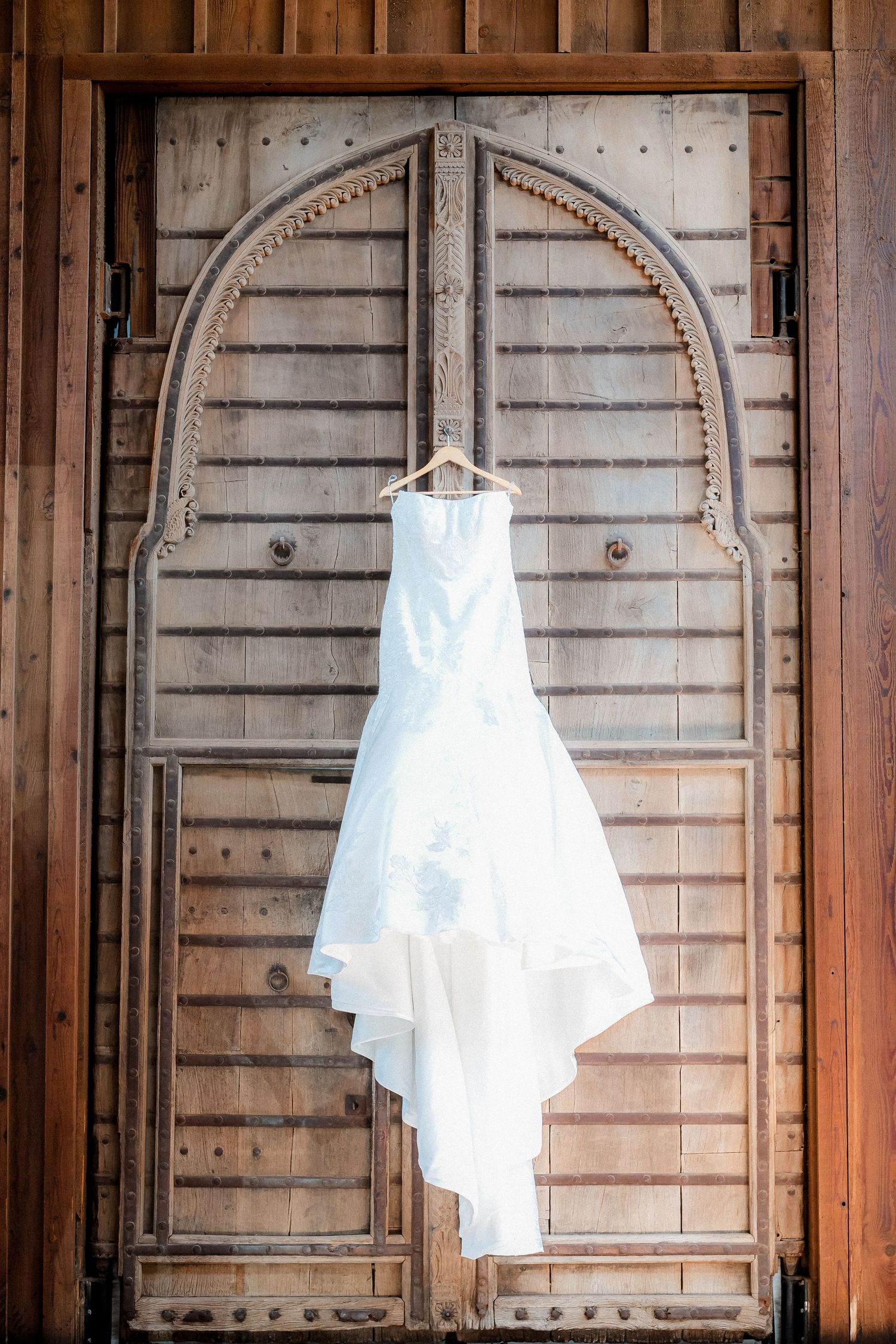 White wedding dress hanging on wooden arched door showcasing its beautiful train and details.