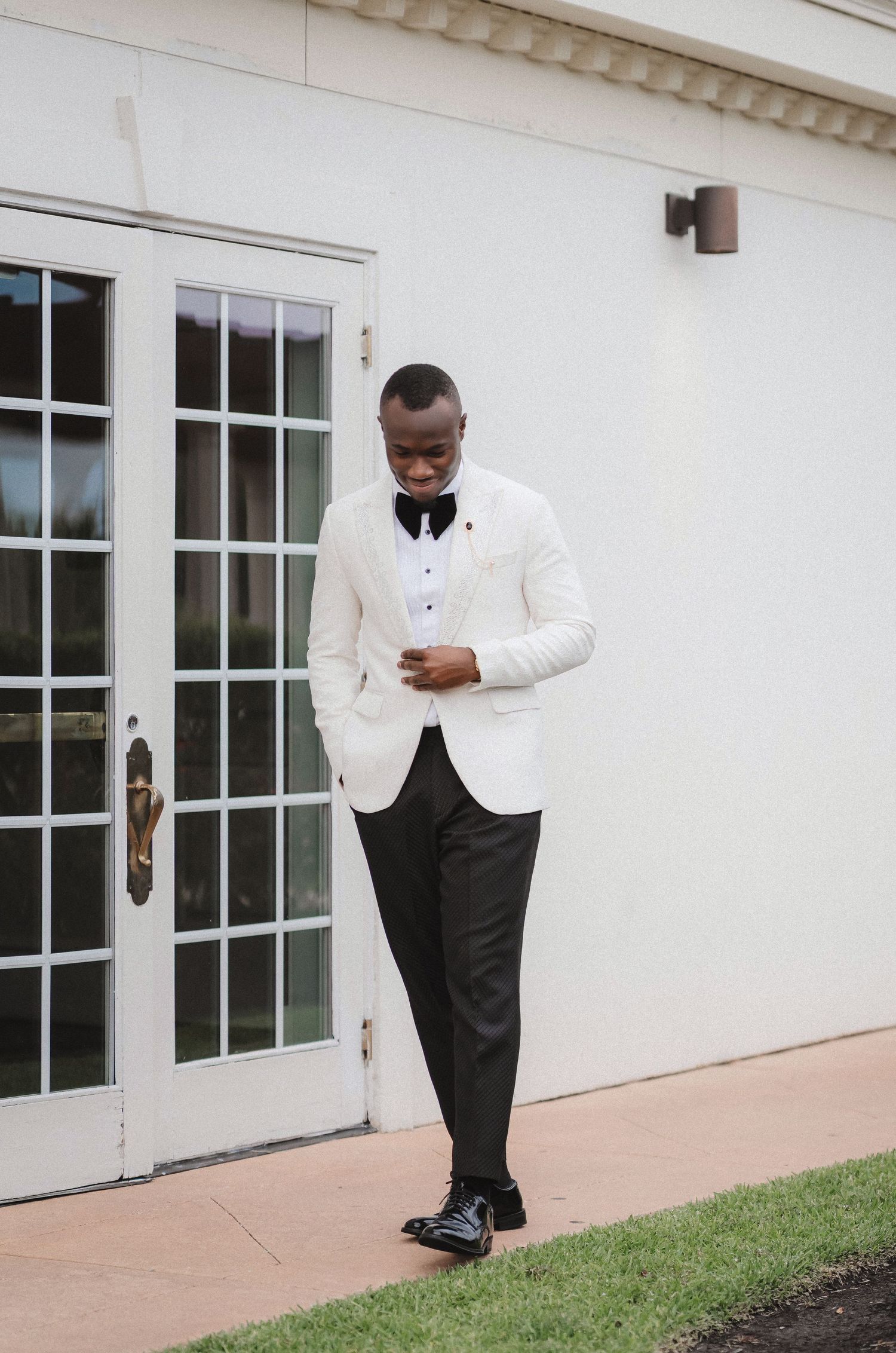 Elegant groom in white tuxedo jacket and black bow tie standing by French doors.