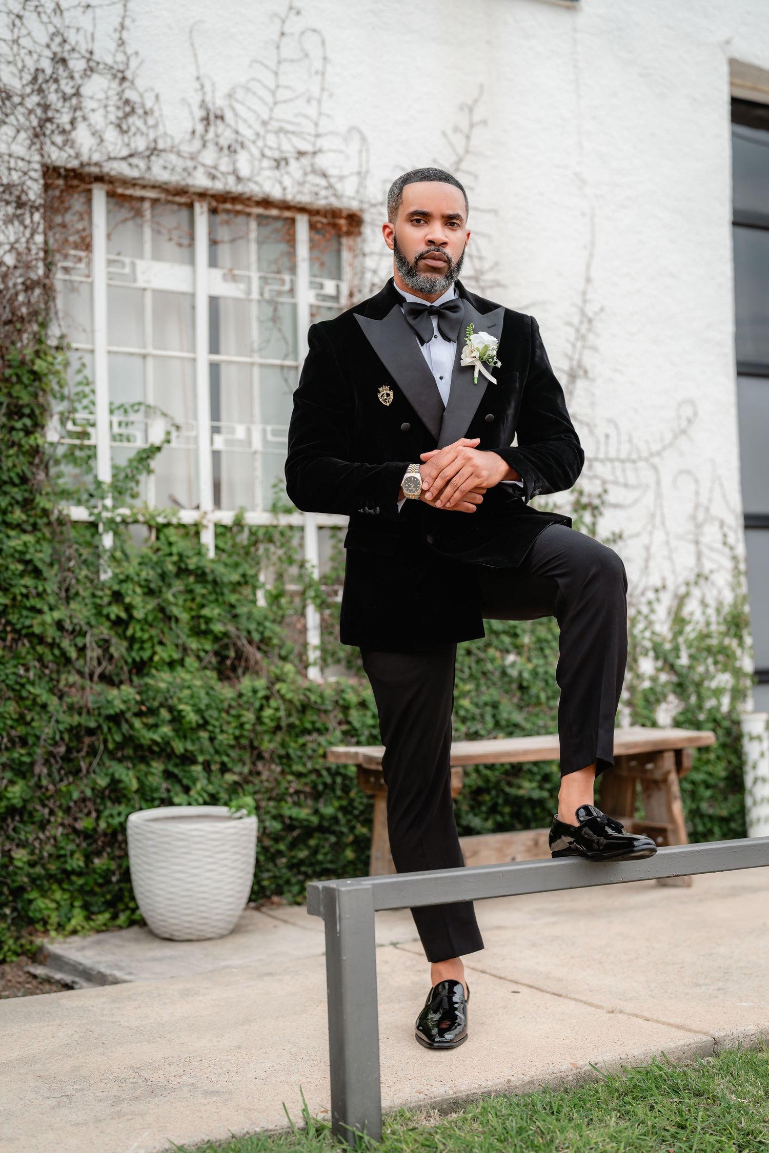 Stylish groom in black velvet tuxedo poses on concrete bench in garden setting.