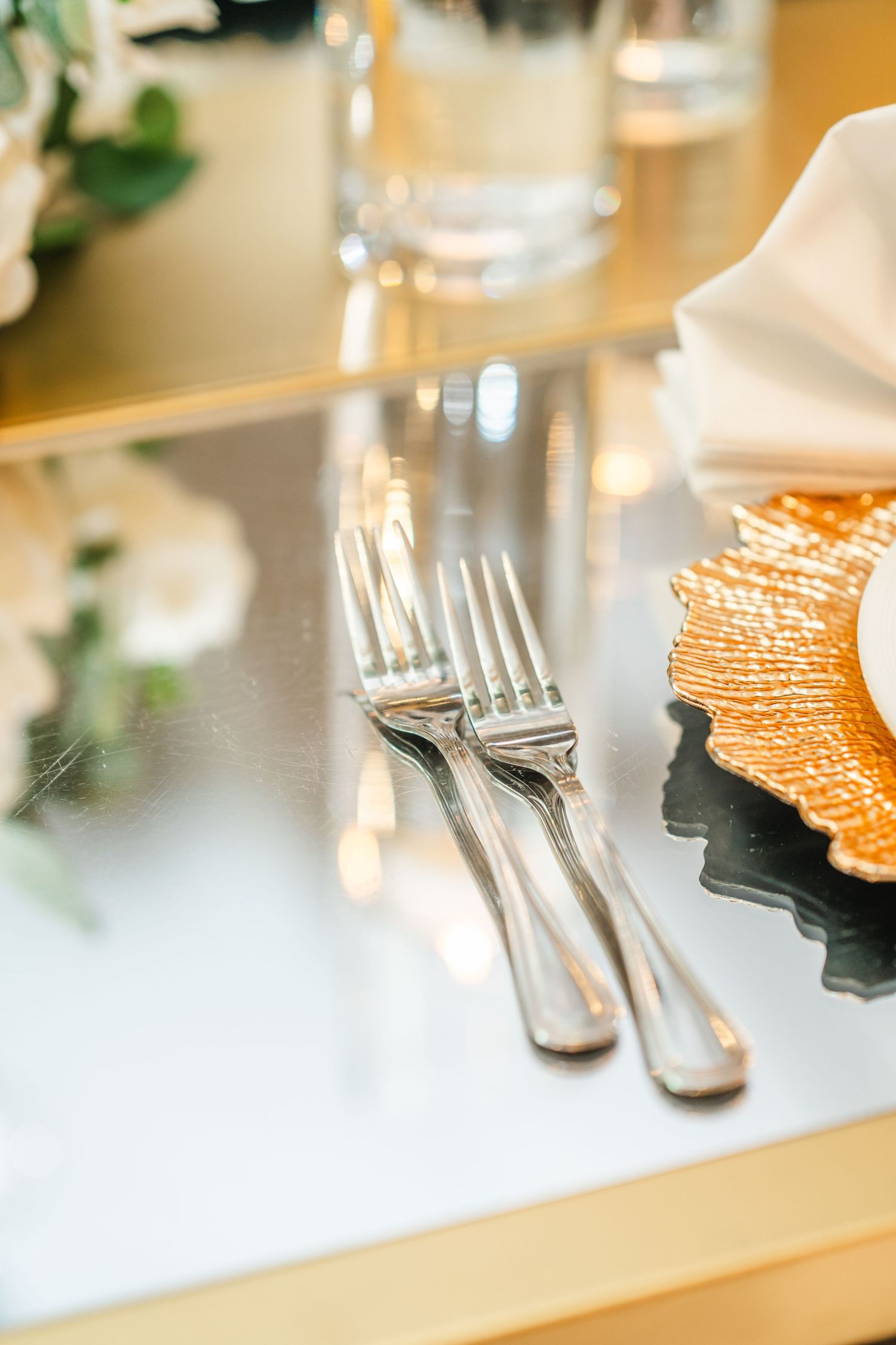Silver forks rest on a reflective surface next to a partially visible plate in an elegant table setting.