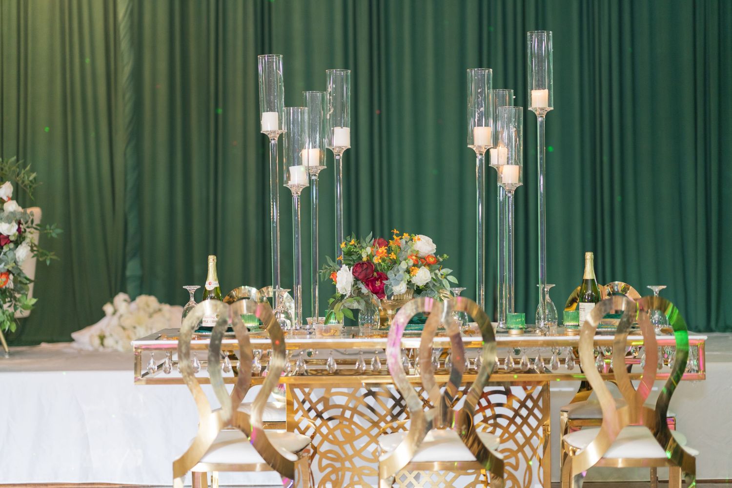 Elegant wedding reception table setup with crystal chandeliers and gold chairs against a green backdrop.