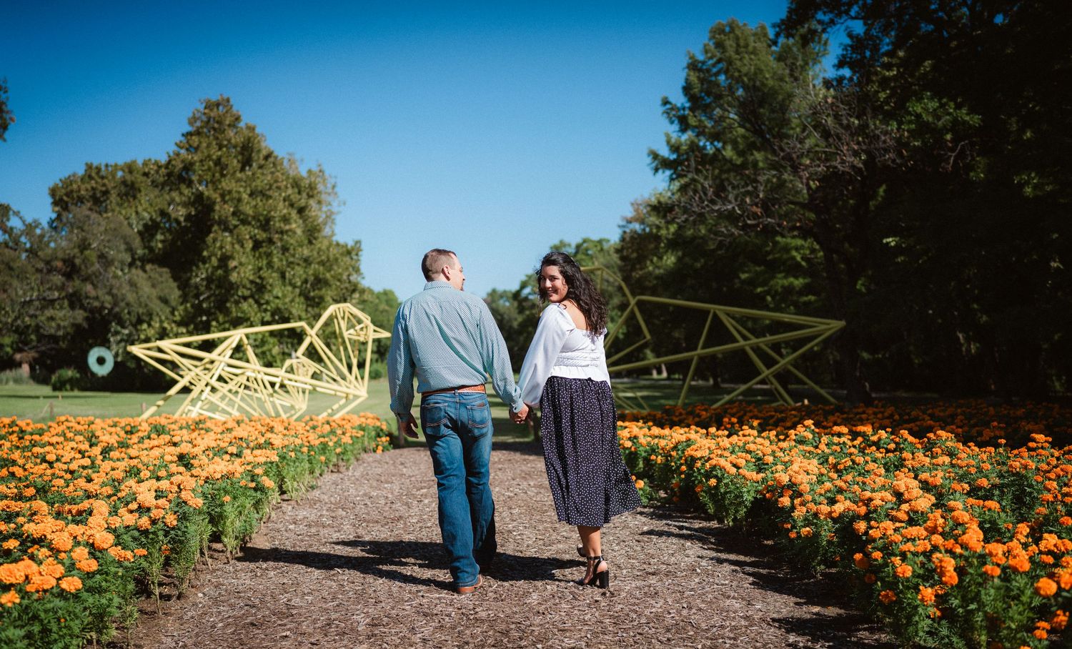 A couple walks along a garden path through vibrant orange marigold flowers on a sunny day.