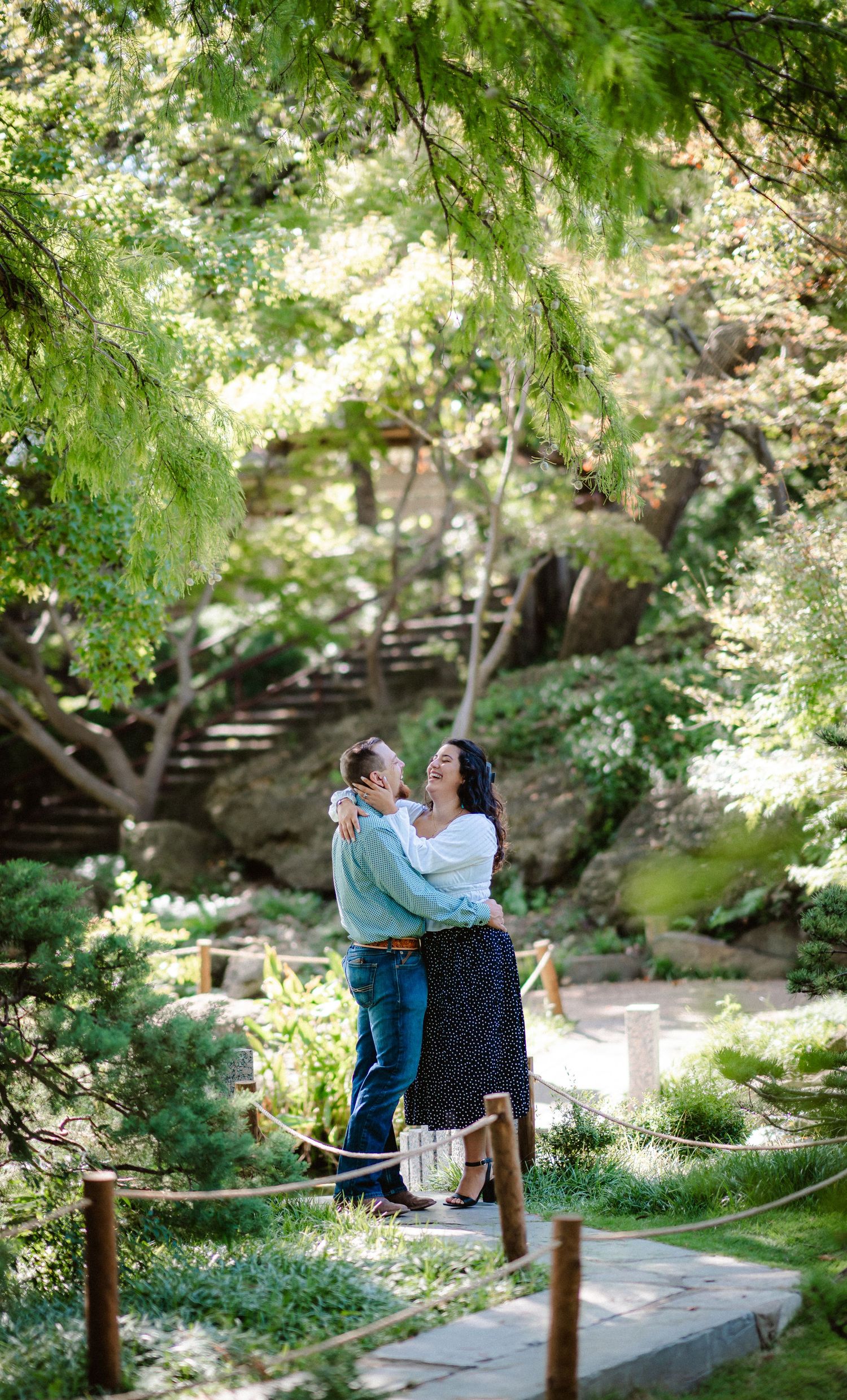 A couple embraces while standing on a stone path surrounded by lush green foliage in a peaceful garden setting.