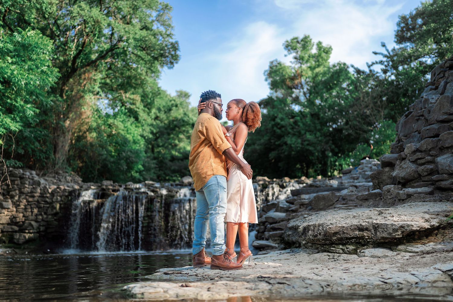 A couple stands together near a scenic waterfall surrounded by lush greenery and rocky terrain.