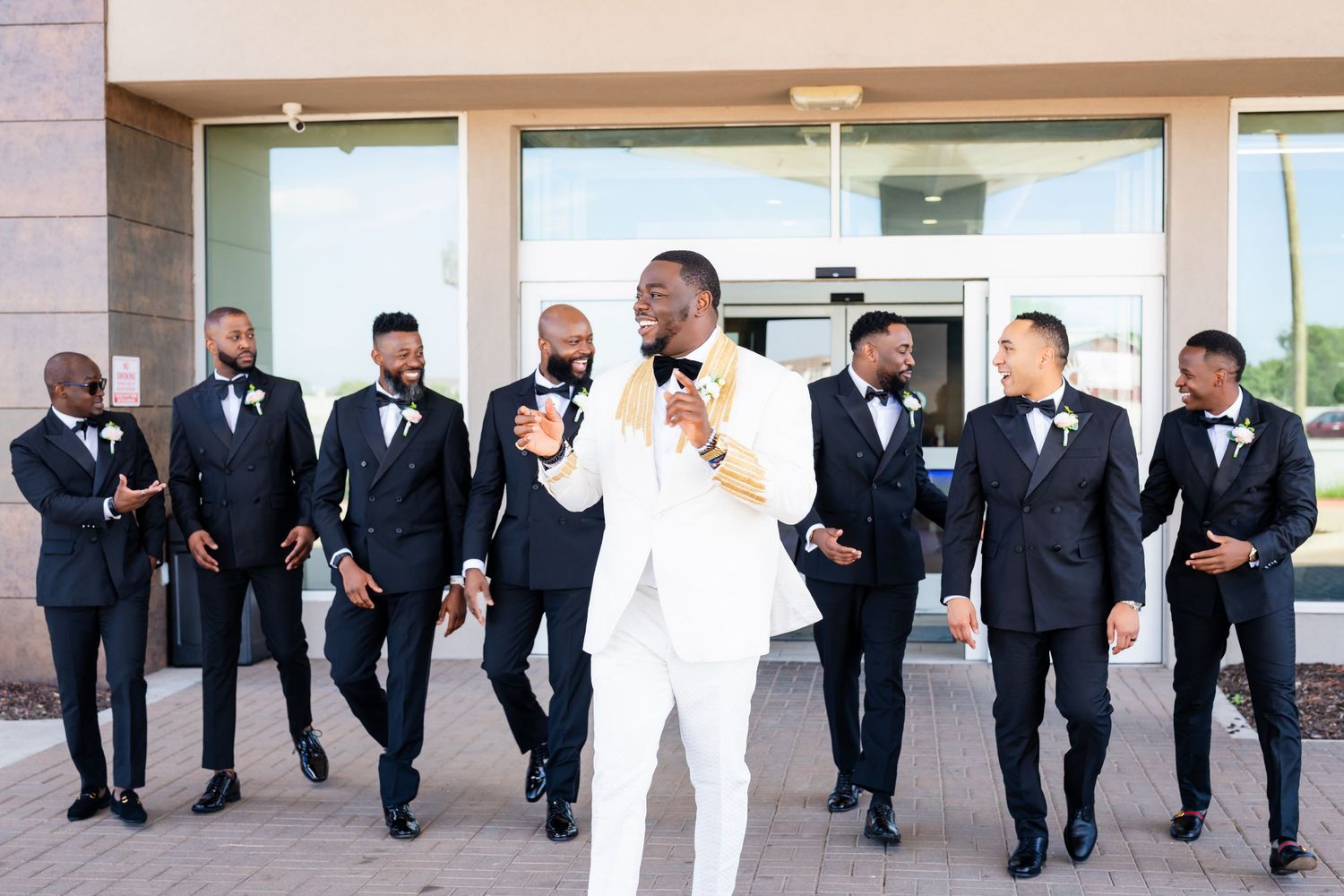 Wedding party in black suits and white tuxedo line up along a modern building exterior.