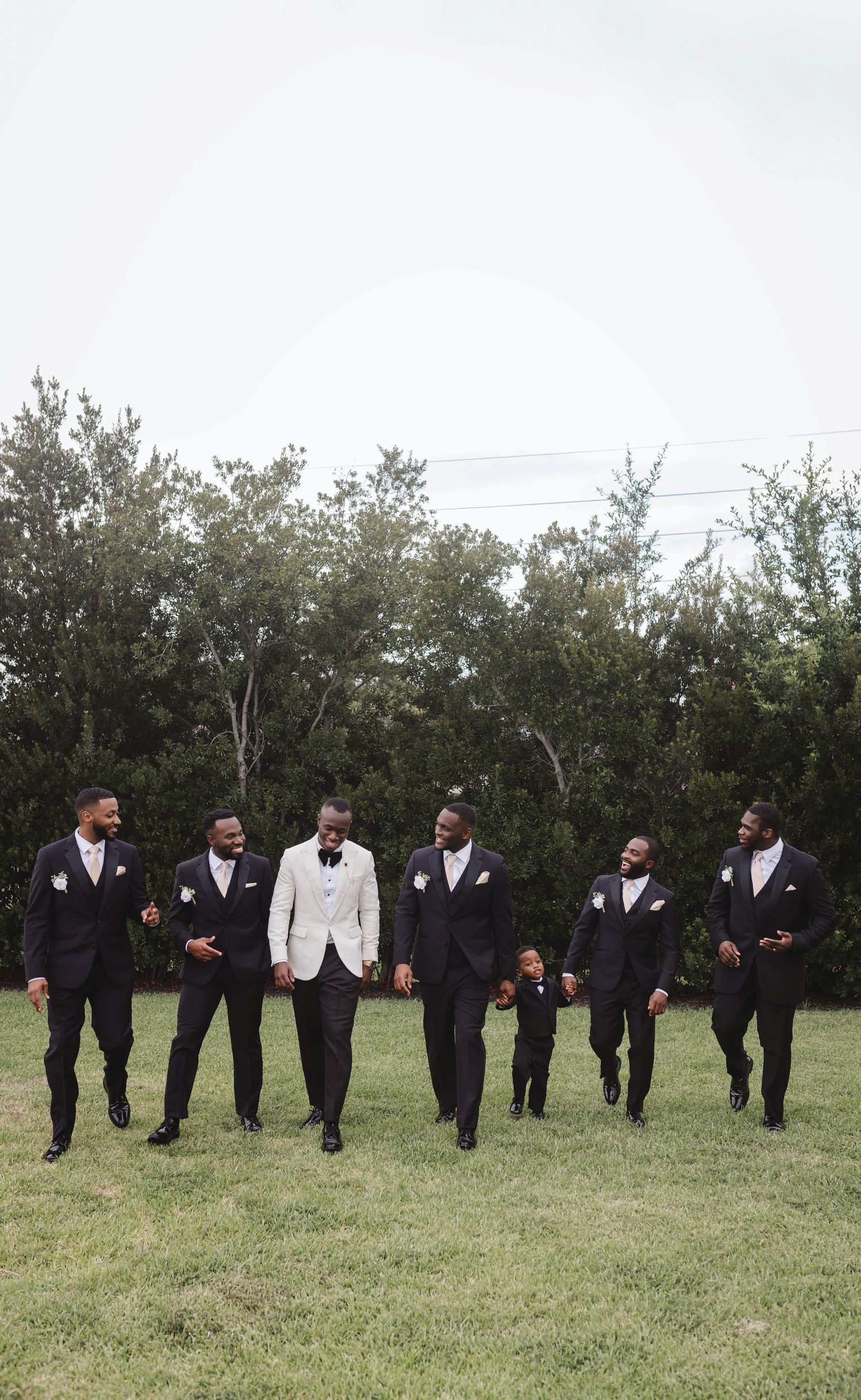 Groomsmen in black suits and one in white walk together on grass with trees in background.