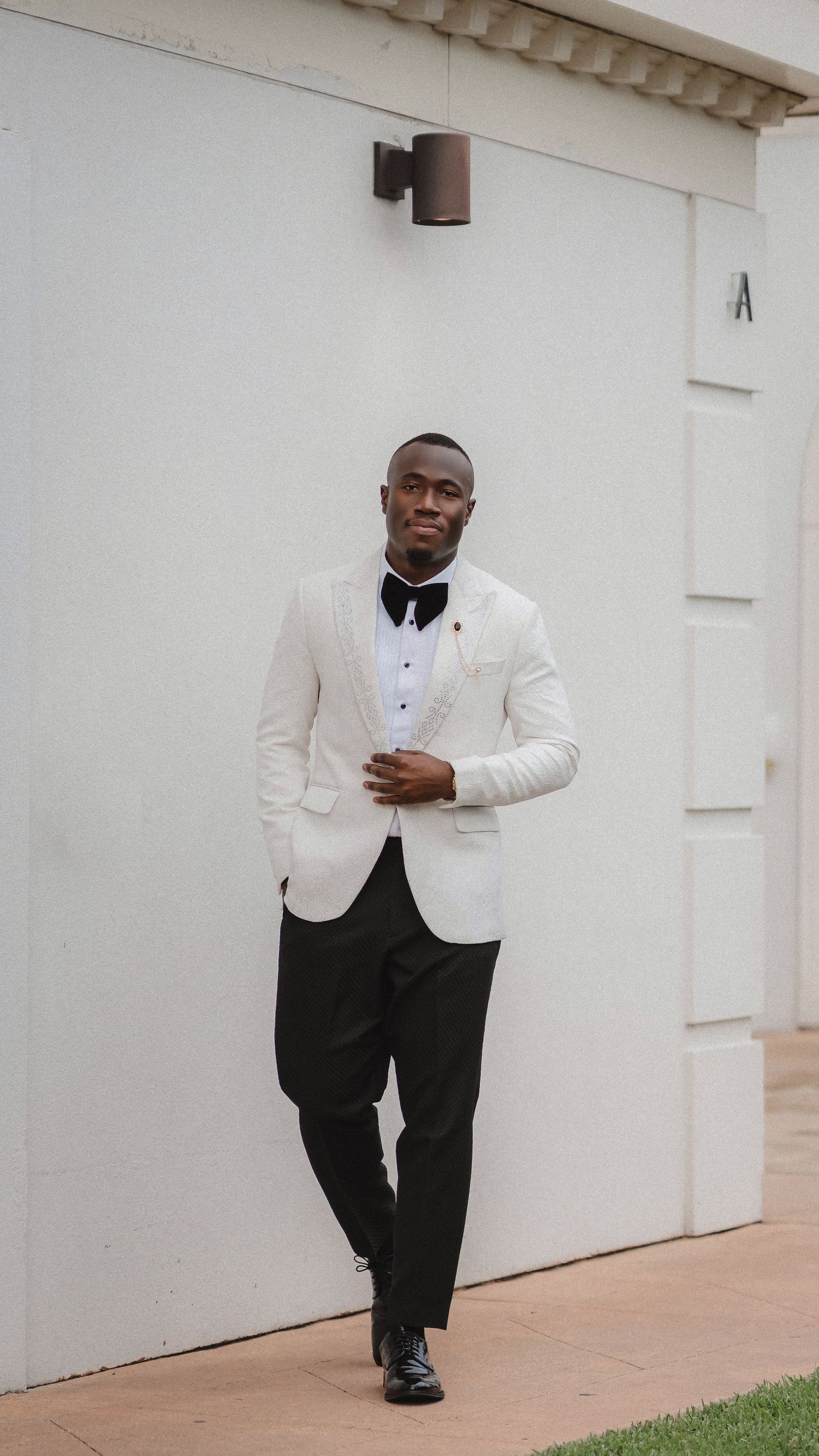 Stylish groom poses against white wall wearing classic white dinner jacket and black bow tie.