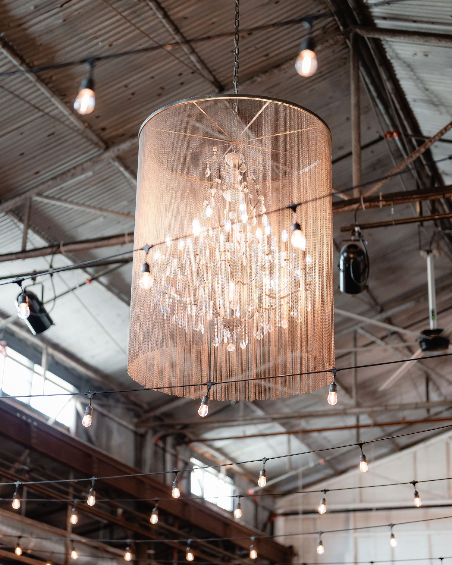 Elegant crystal chandeliers and string lights illuminate an industrial-style wedding venue ceiling.
