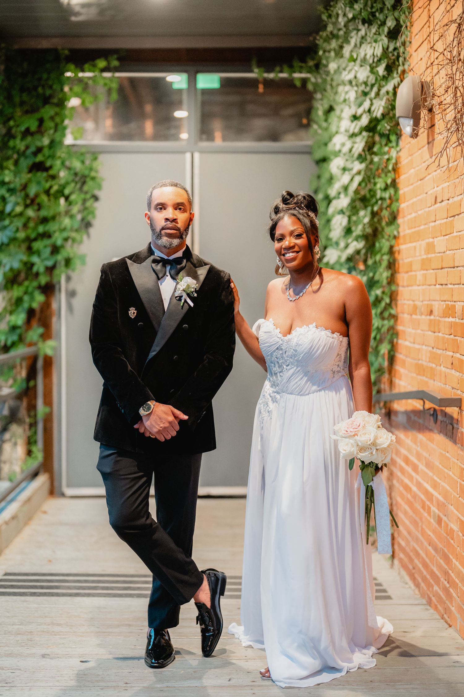 Playful wedding couple posing along a brick wall walkway with greenery and modern architecture.