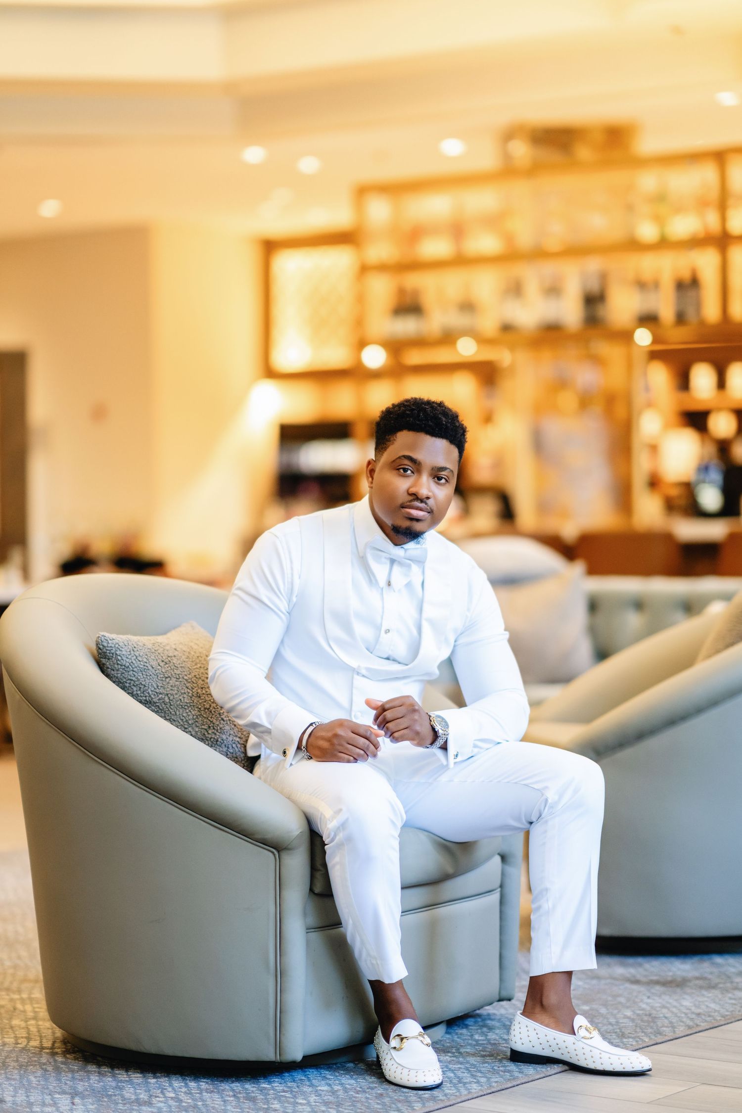 A person in an all-white outfit sits elegantly on a modern grey lounge chair in an upscale venue.