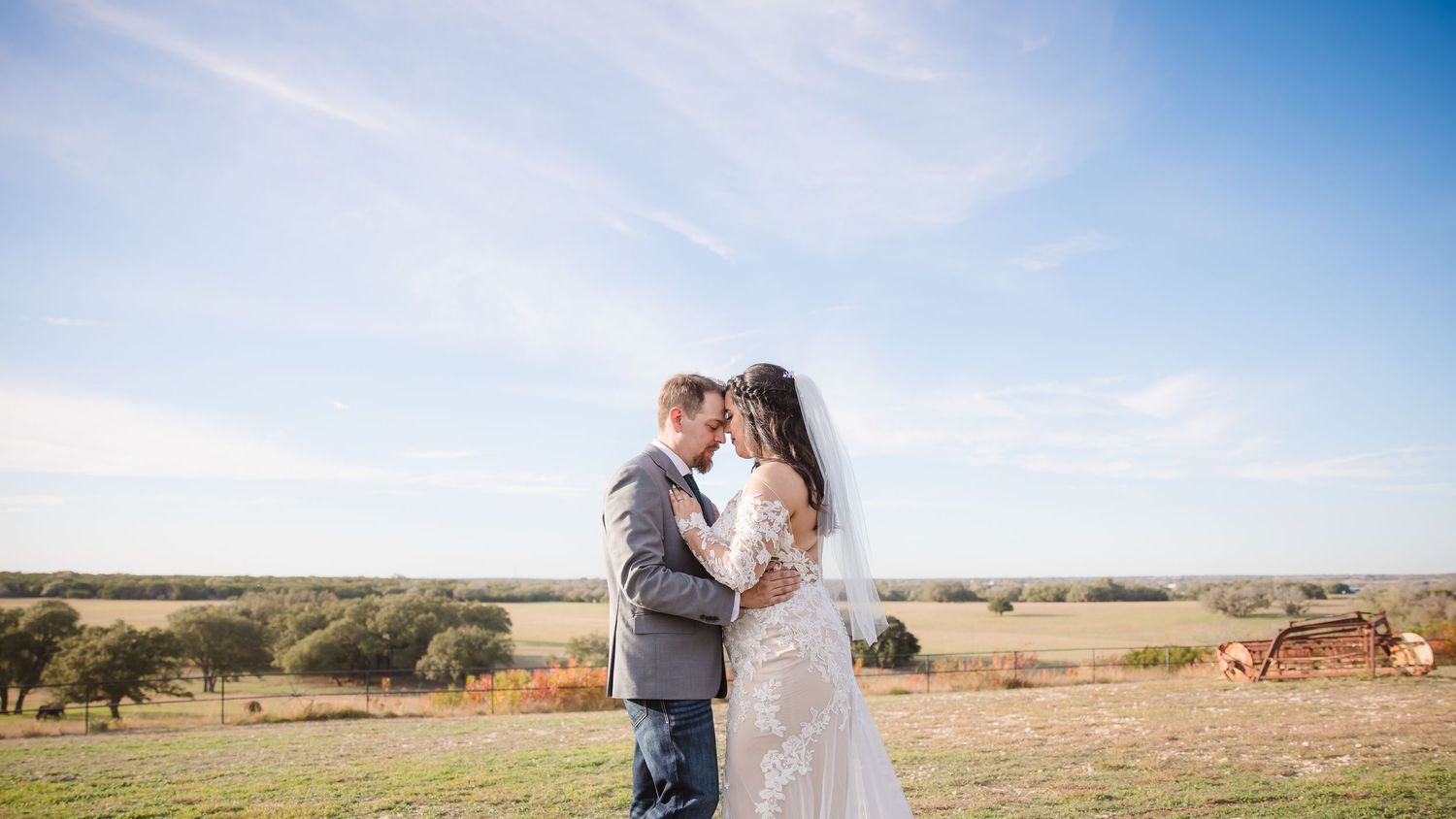 A couple embraces in an open field with mountains and blue sky in the background during their outdoor wedding shoot.