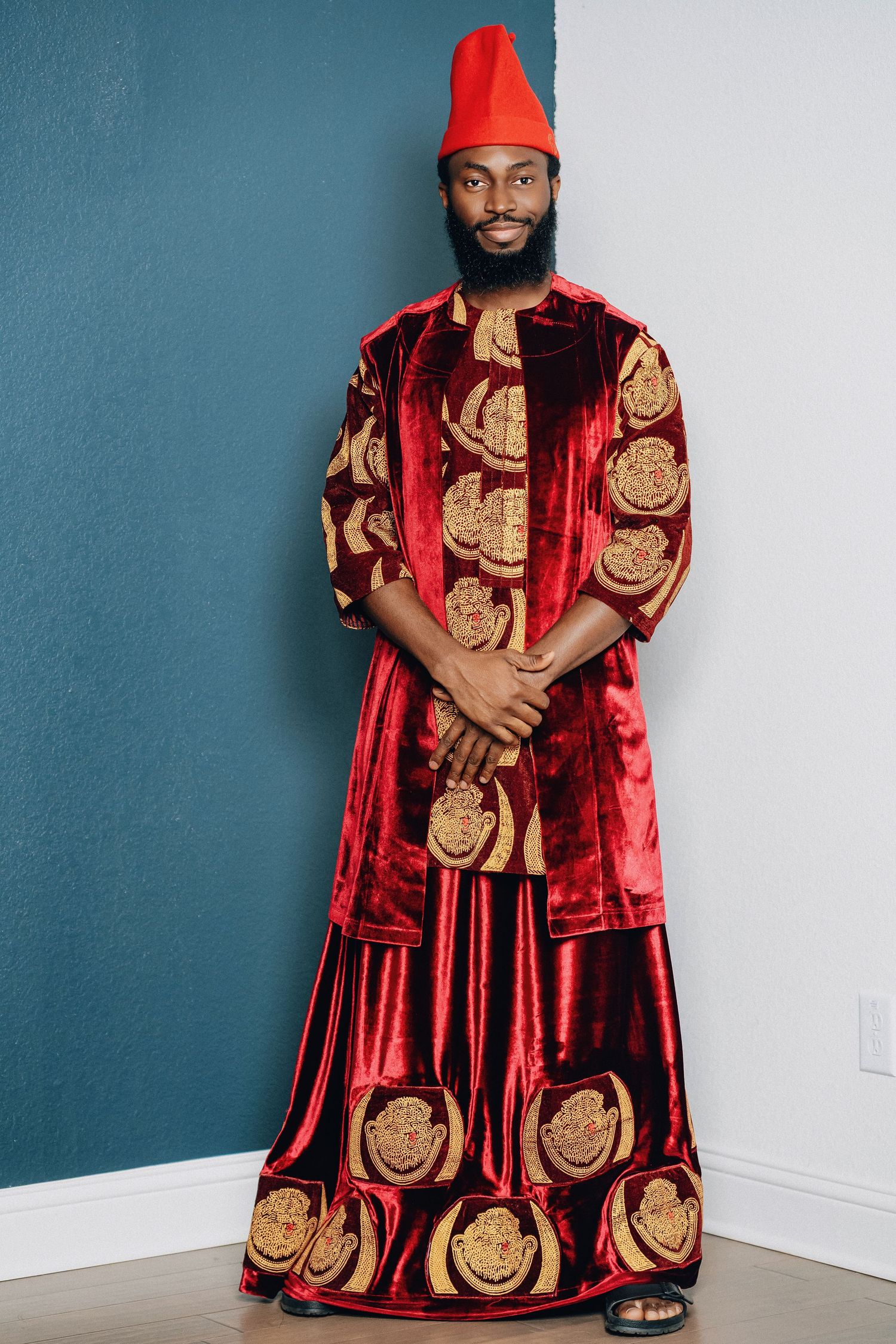 A person wearing a traditional red and gold patterned Nigerian agbada with matching cap against a teal wall.