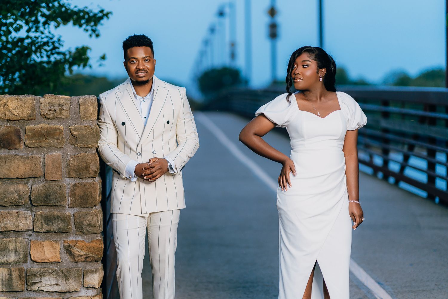 A couple in coordinating white outfits poses together on a boardwalk during a romantic outdoor photoshoot.