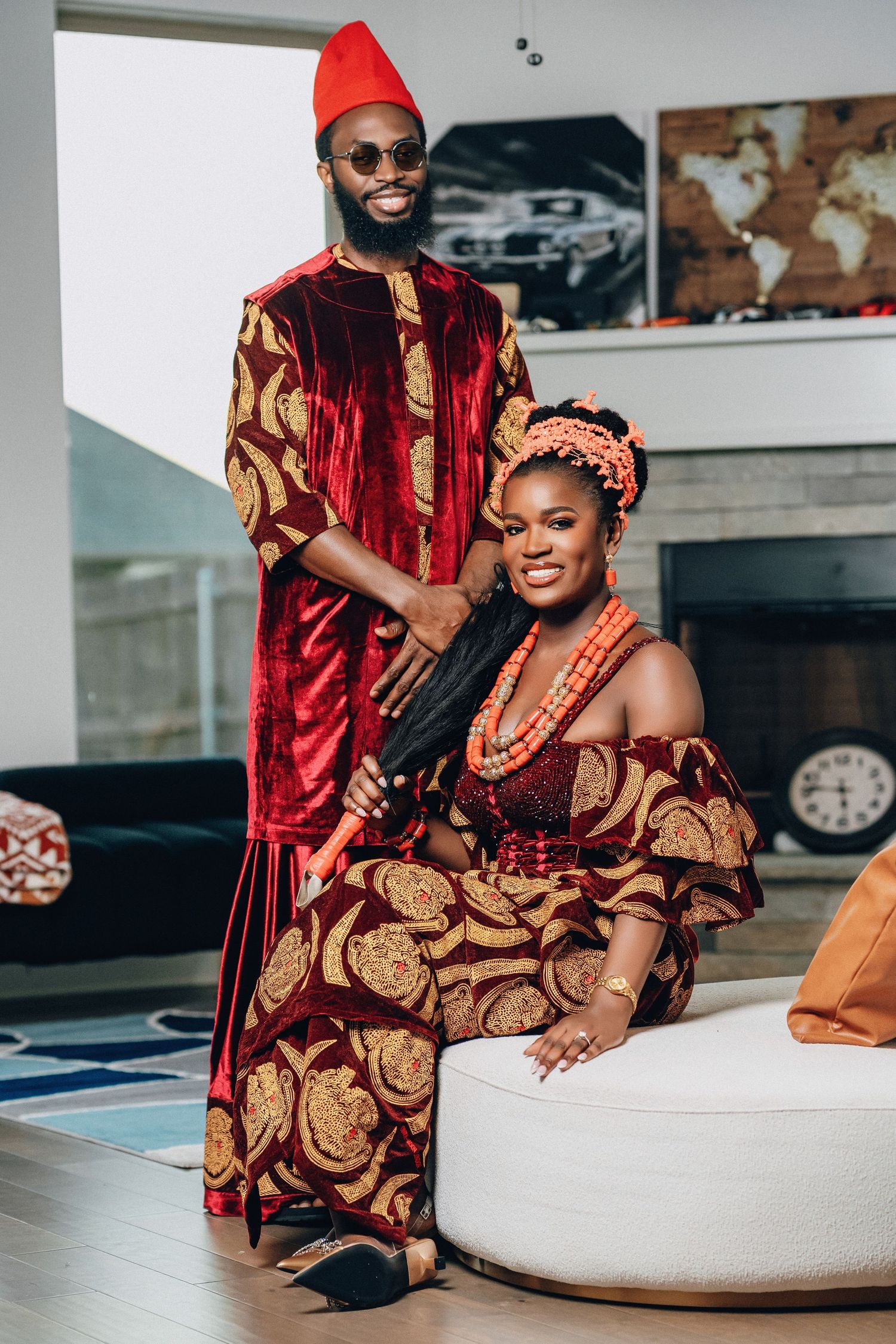 A couple in traditional Nigerian attire poses together on a white couch wearing red and gold matching outfits.