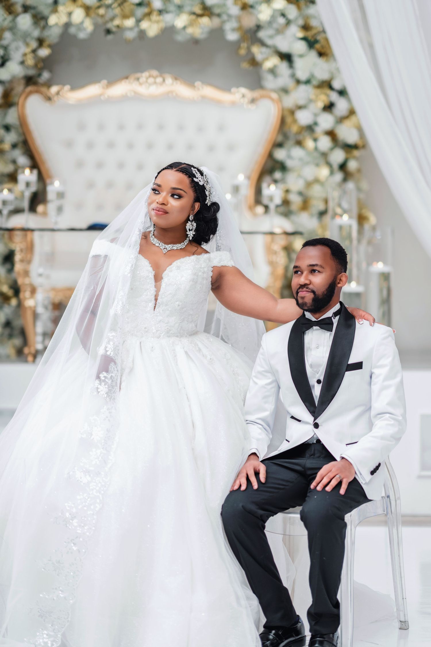Newlyweds pose elegantly in white wedding attire against a luxurious floral backdrop.